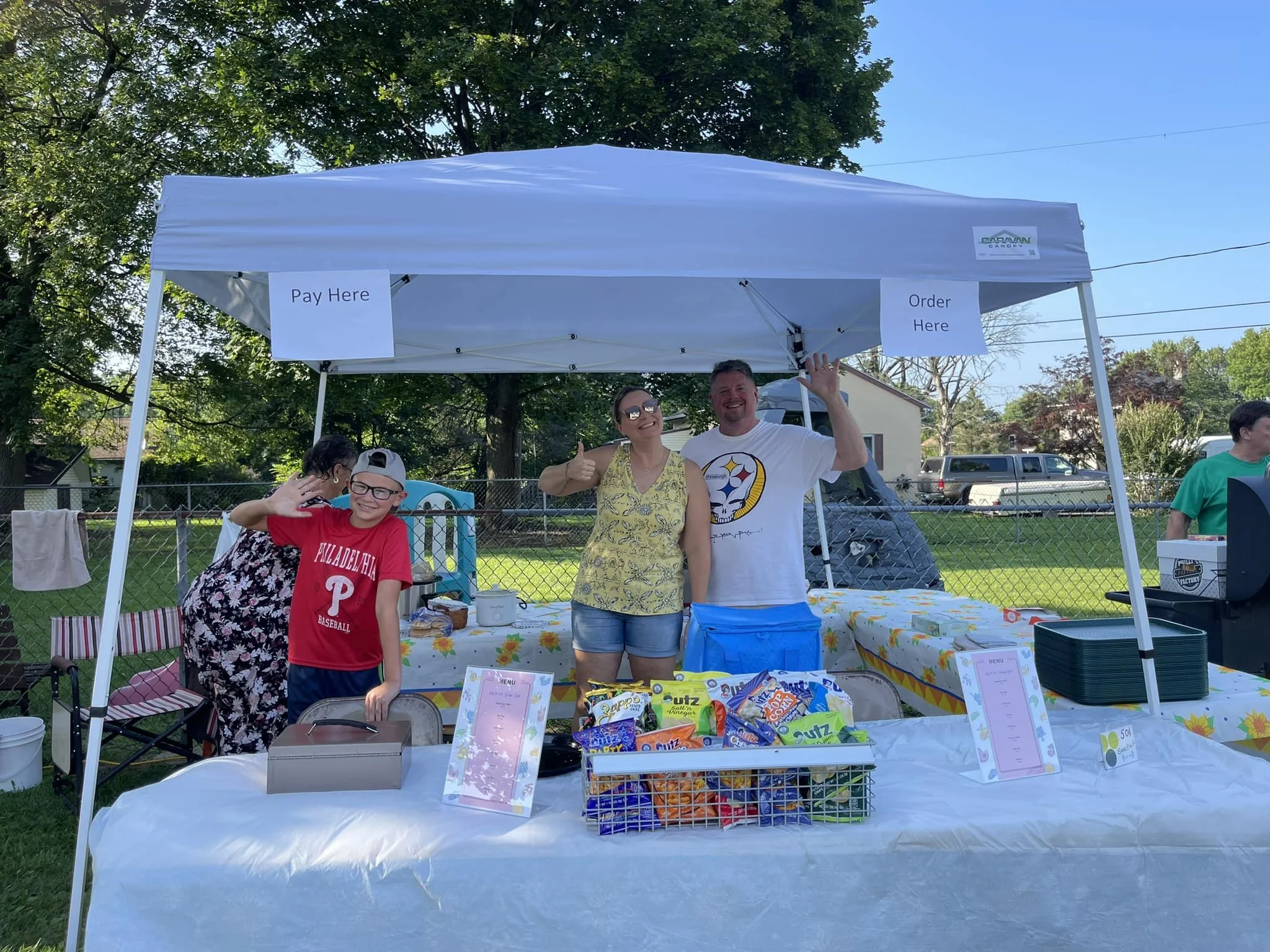 A group of people standing behind a table at an outdoor event with a white tent overhead. The table has various snacks and menus, and there are signs that say 'Pay Here' and 'Order Here.' Trees and houses are visible in the background.