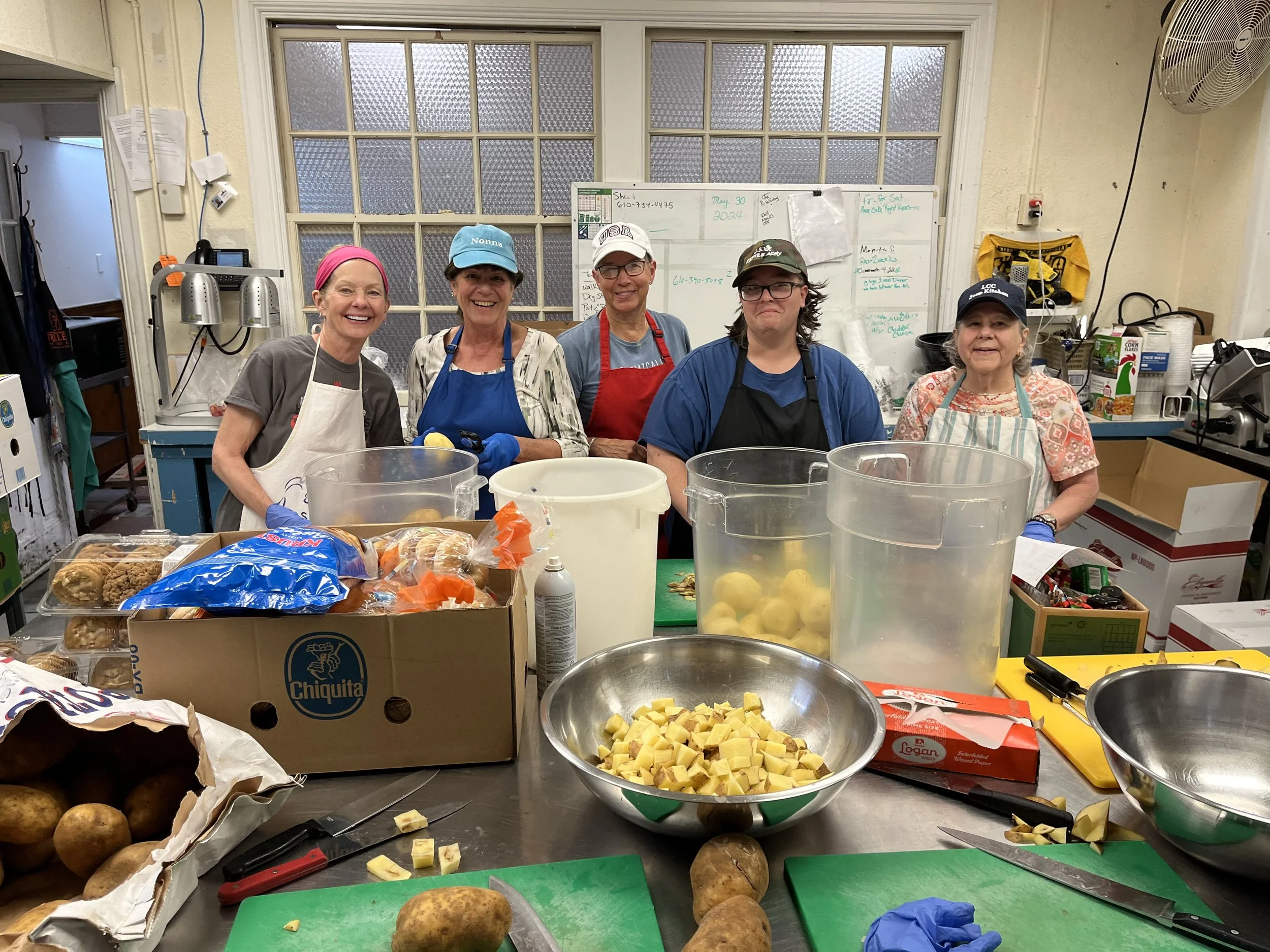 Group of five smiling women wearing aprons and hats standing behind a kitchen counter full of ingredients and utensils.