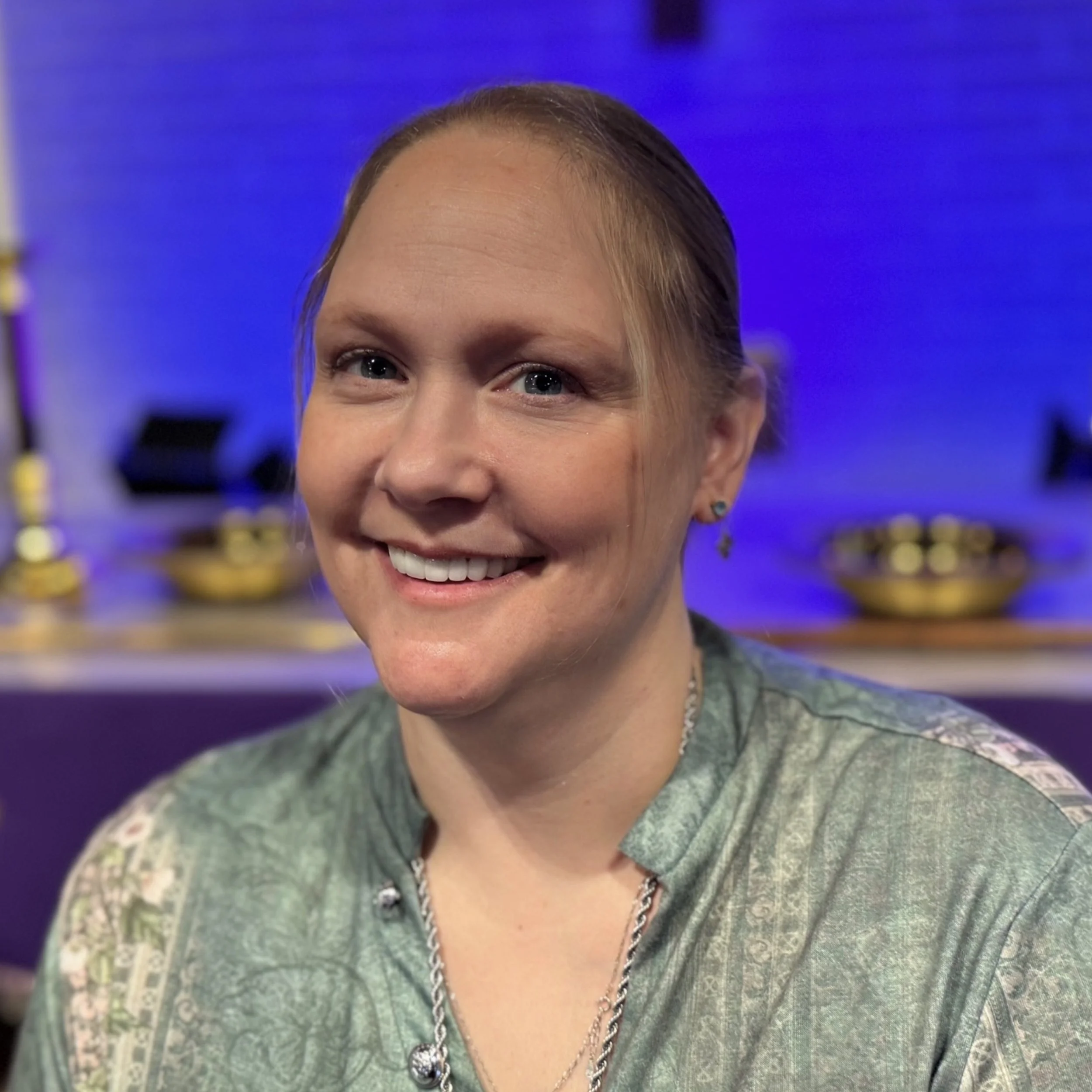Close-up of a smiling woman with blue eyes and light brown hair, wearing a green patterned top and jewelry, in front of a blue and purple background.