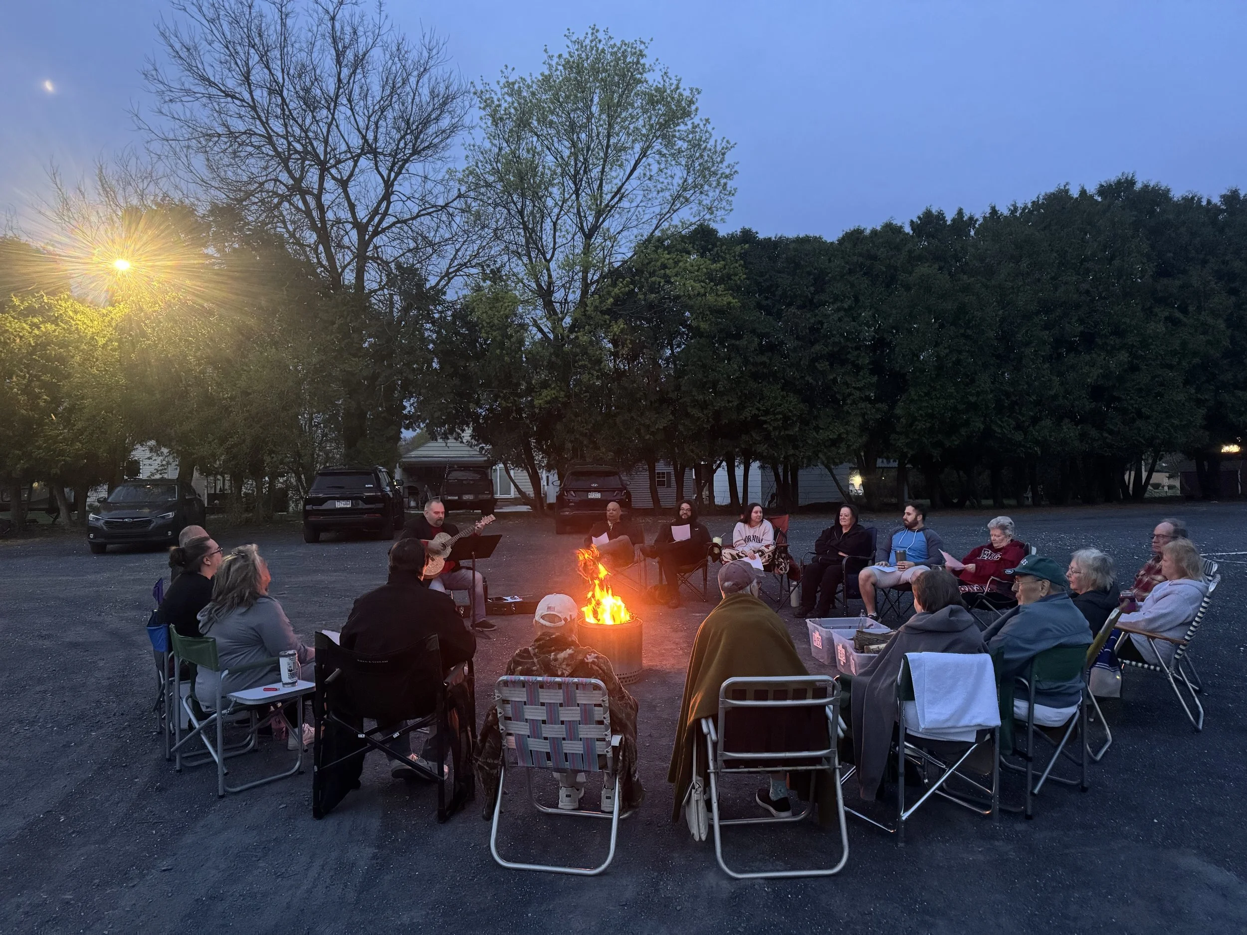 People sitting around a campfire during twilight in a parking lot, with a backdrop of trees and houses, some holding papers, one playing guitar.