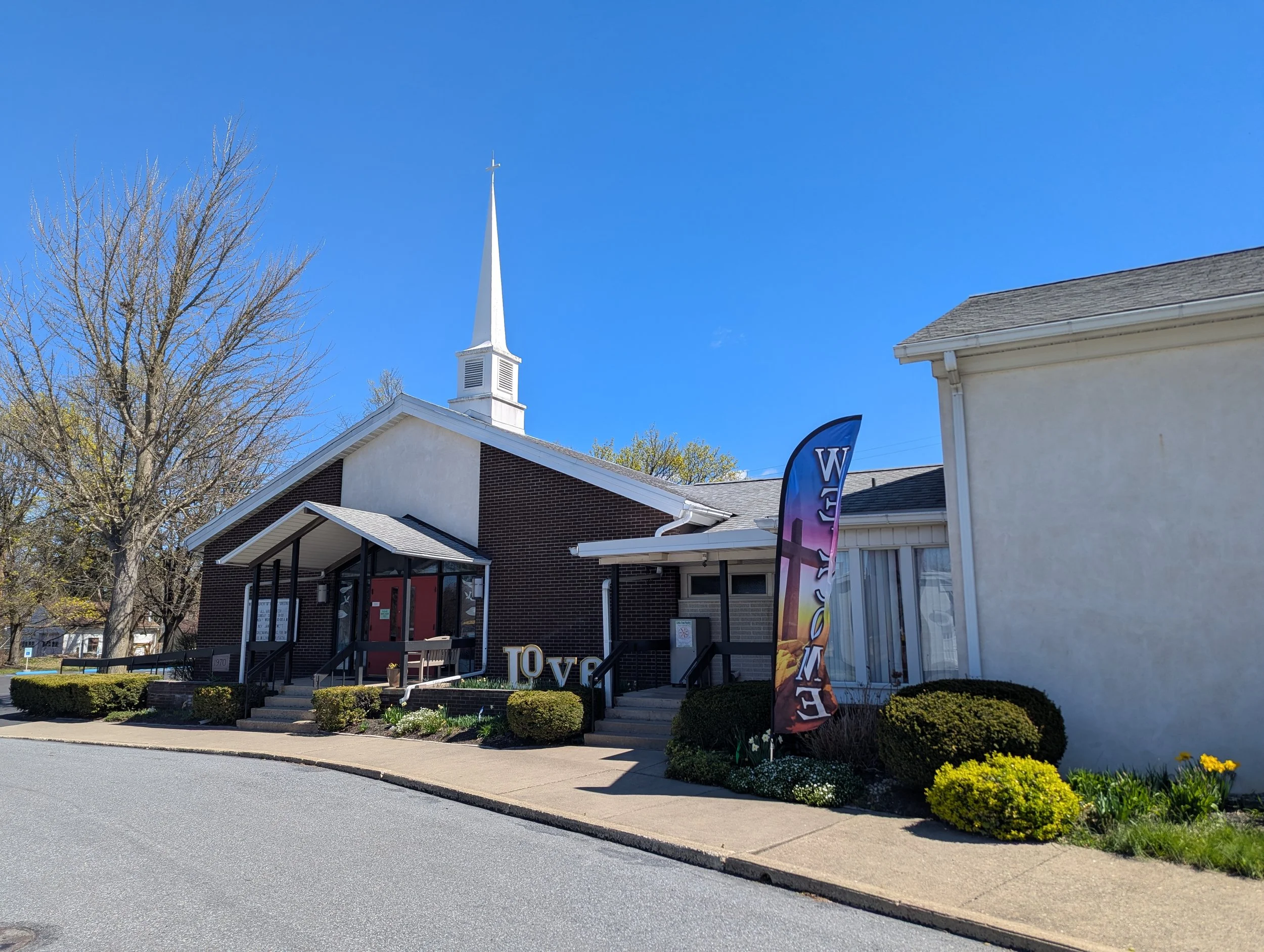 A church with a tall steeple, a small garden with bushes and flowers, a banner with the word 'LOVE', and a sidewalk in front of the building under a clear blue sky.