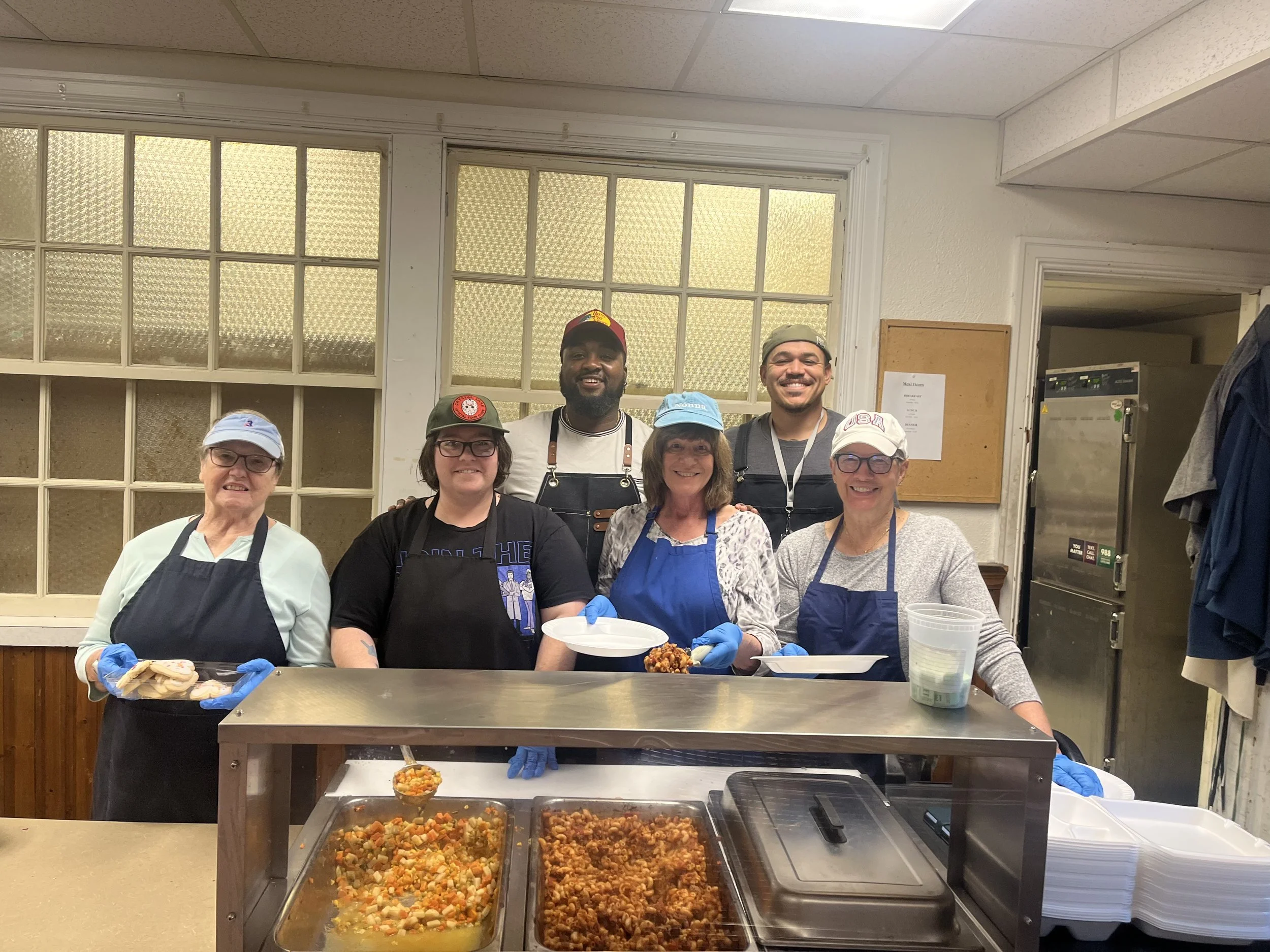 Group of six diverse people in hairnets and aprons standing behind a food service counter with trays of cooked food, smiling at the camera.