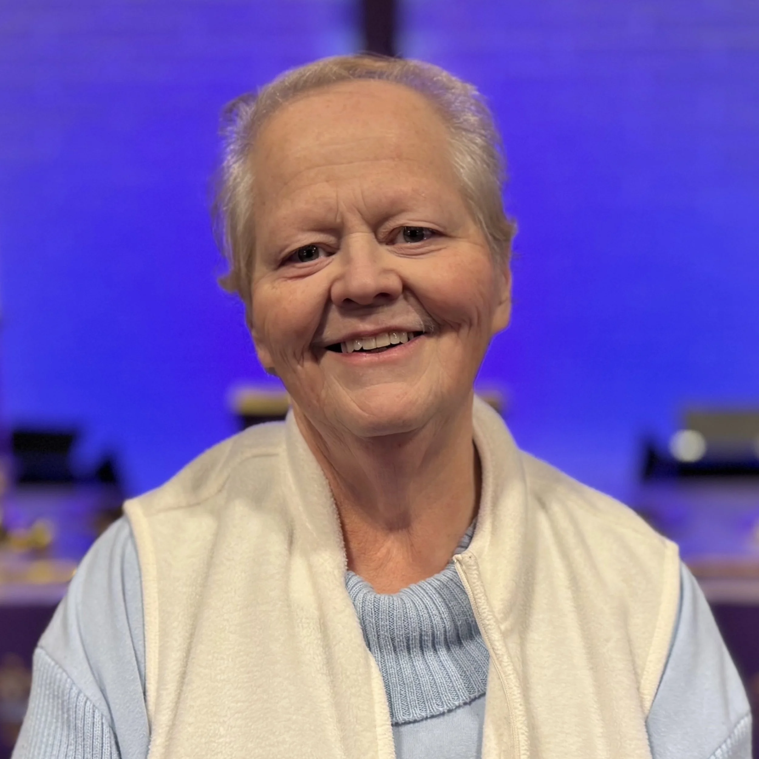 Close-up of smiling elderly woman with gray hair, wearing a beige jacket and a light blue sweater, against a blue background.