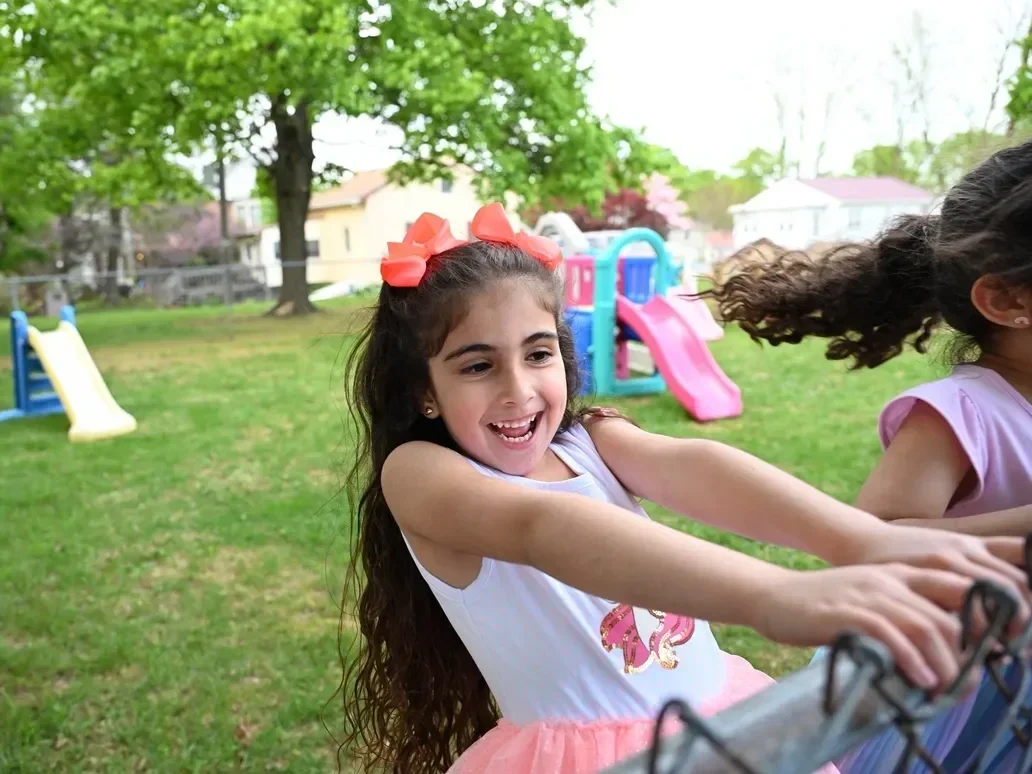 Young girl smiling while playing outdoors on playground equipment with another girl nearby.