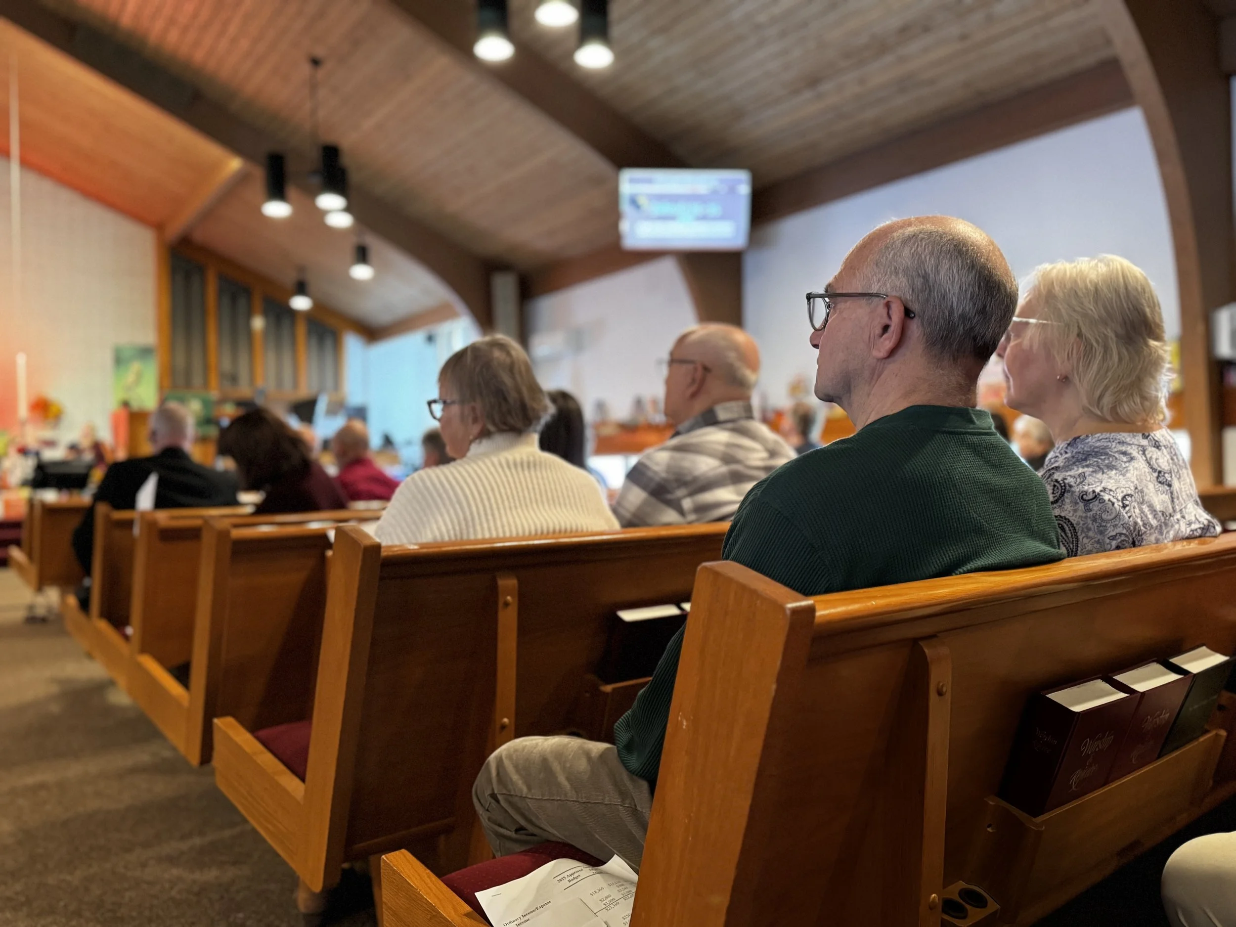People seated in wooden pews inside a church or meeting hall, facing forward. The room has a sloped wooden ceiling with hanging lights and a projection screen on the wall.