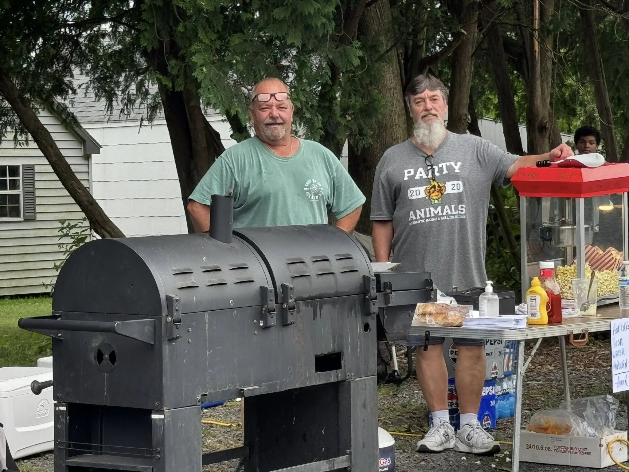 Two smiling men standing behind a grill and a table with food and condiments at an outdoor event, with trees and a house in the background.