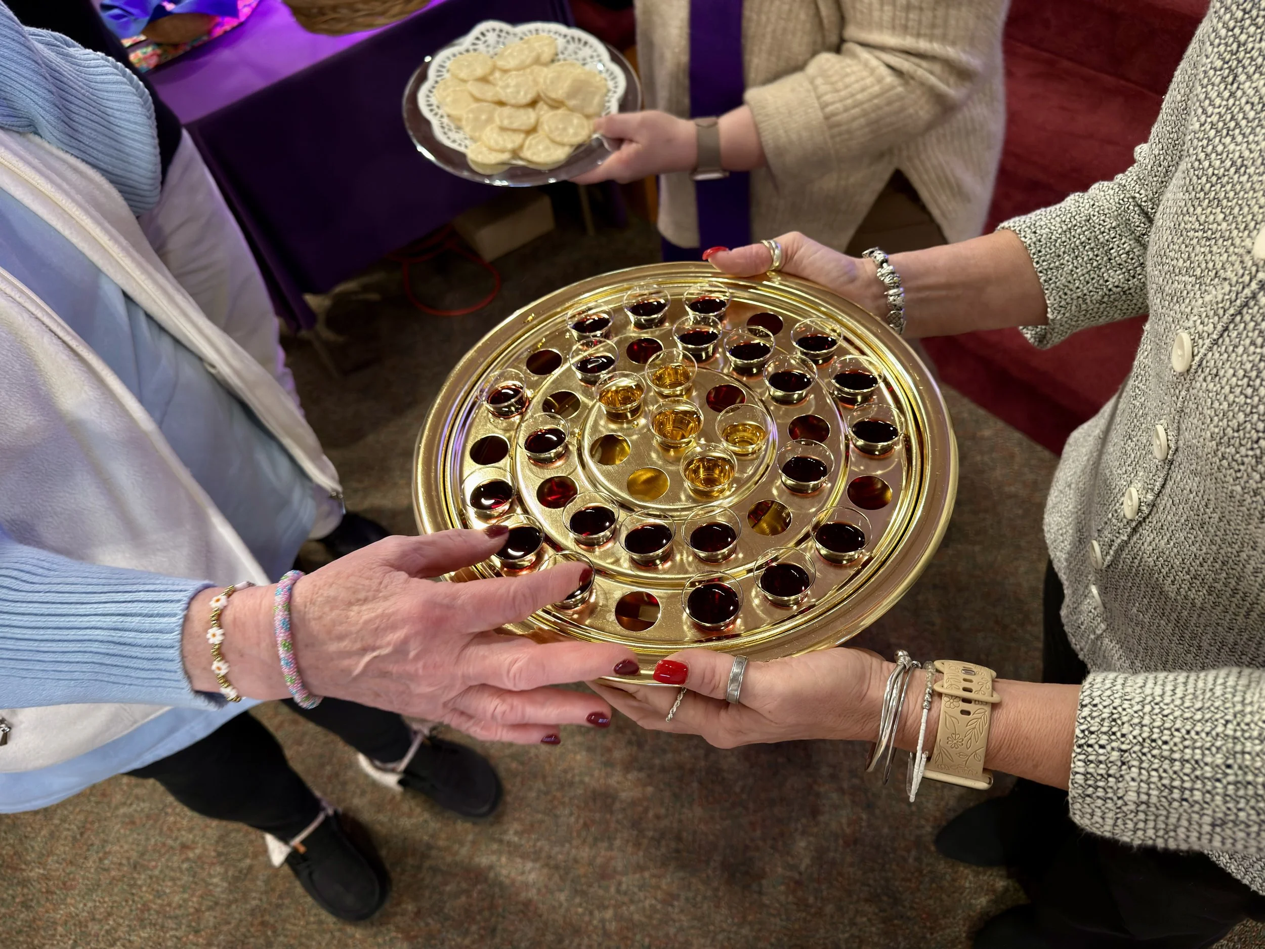 People exchange drinks and snacks at a social gathering. One person holds a tray with small glasses filled with dark-colored liquid, and another person holds a plate of small snacks or cookies.
