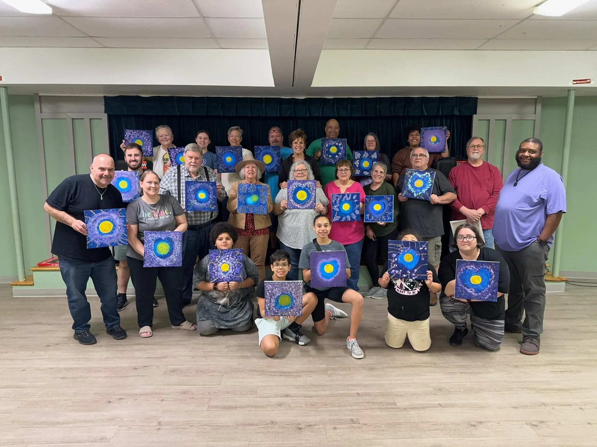 Group of smiling people of various ages holding up paintings of a blue background with a yellow circle in the center, standing in a room with a stage and dark curtains.