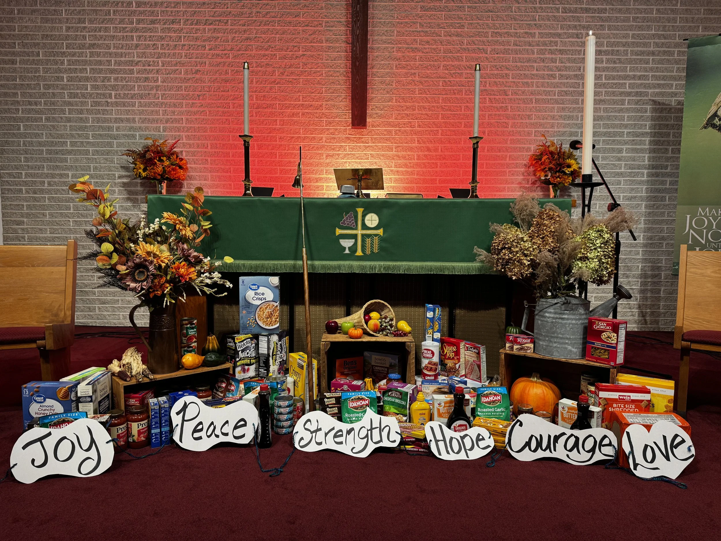 A church altar decorated with fall flowers and pumpkins, with a green cloth featuring religious symbols. In front, there are various food items, canned goods, and signs with words like 'Joy,' 'Peace,' 'Strength,' 'Hope,' 'Courage,' and 'Love' arrange