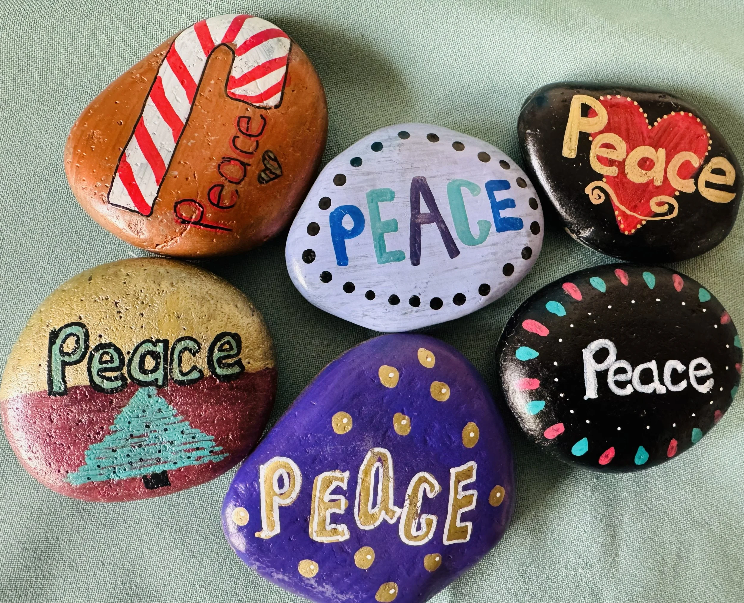 Painted rocks with words and designs, including the words "peace" and "peace" on some, and other words like "hope" and "peace" on others, with colorful patterns and decorative elements, placed on a green fabric background.
