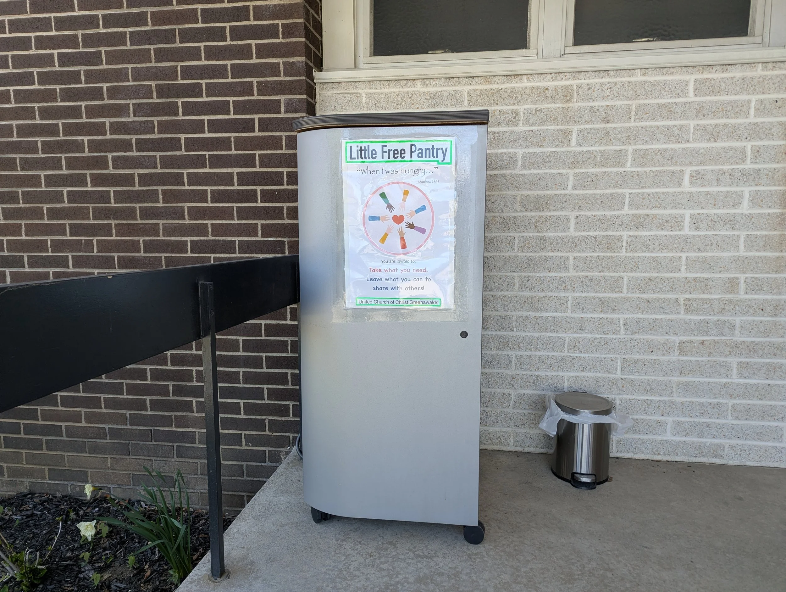 A gray bin labeled 'Little Free Pantry' located outside against a brick and vinyl siding wall. The sign invites people to take what they need, leave what they can, and share with others. There is a plant with white flowers, a black bench, and a silver trash can nearby.