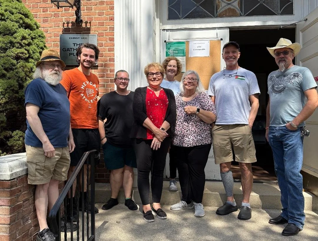 Group of nine people standing together outside in front of a church entrance, smiling for the photo. The group includes men and women of various ages, some wearing casual clothing and hats.