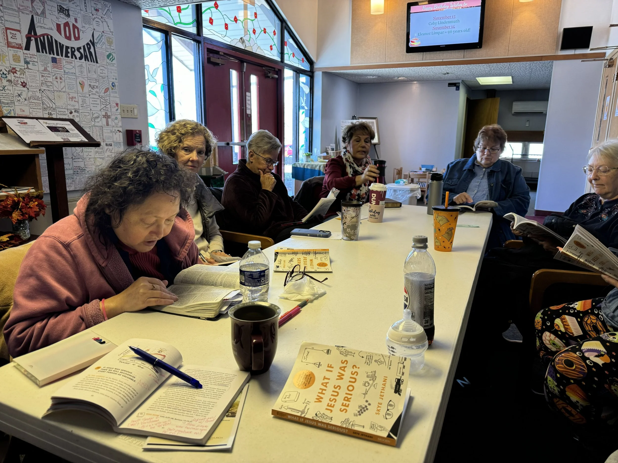 A group of seven women sitting around a long table in a room, with books, papers, and cups in front of them, engaged in reading or discussion. The room has large windows with stained glass designs and a display board with a 10th anniversary poster on the wall.