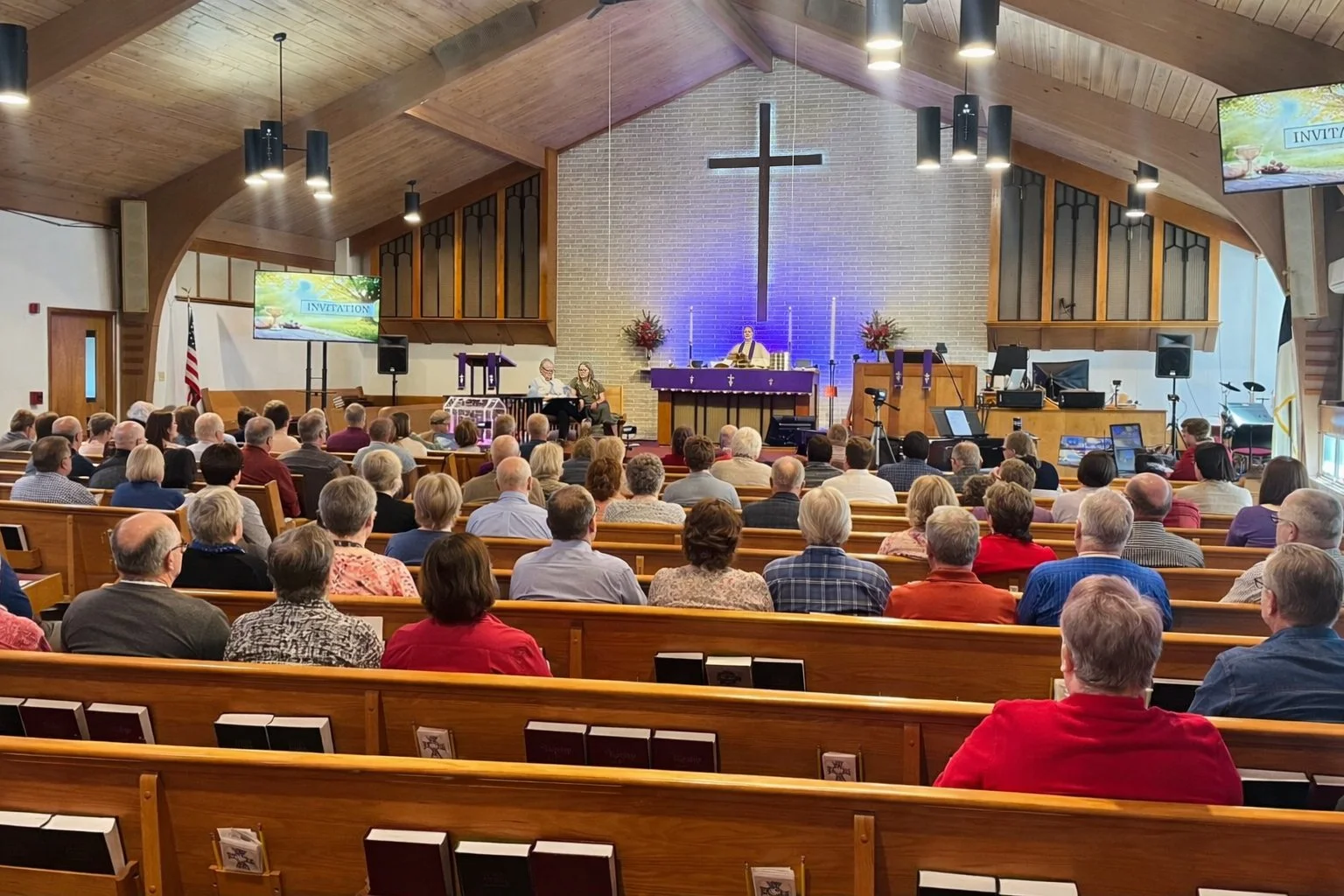 Inside a church during a service or event, with a congregation seated facing the altar. The altar has a large cross on brick wall background, and is decorated with flowers and candles. Attendees are listening to a speaker at the pulpit and a group seated at a table on the stage. Large screens display the word 'INVITATION'. The church has wooden ceilings and pews, with side windows and screens.