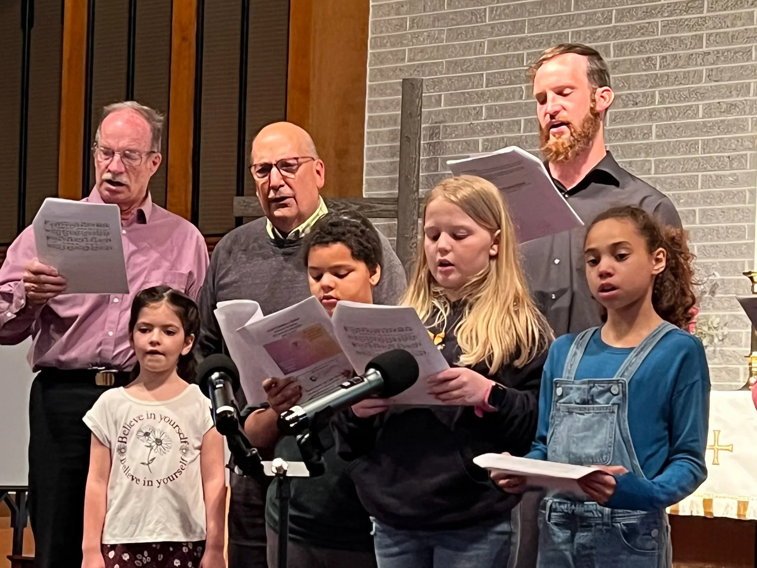 A group of people singing from songbooks during a church service, with three children and three adults visible, standing in front of a brick wall.