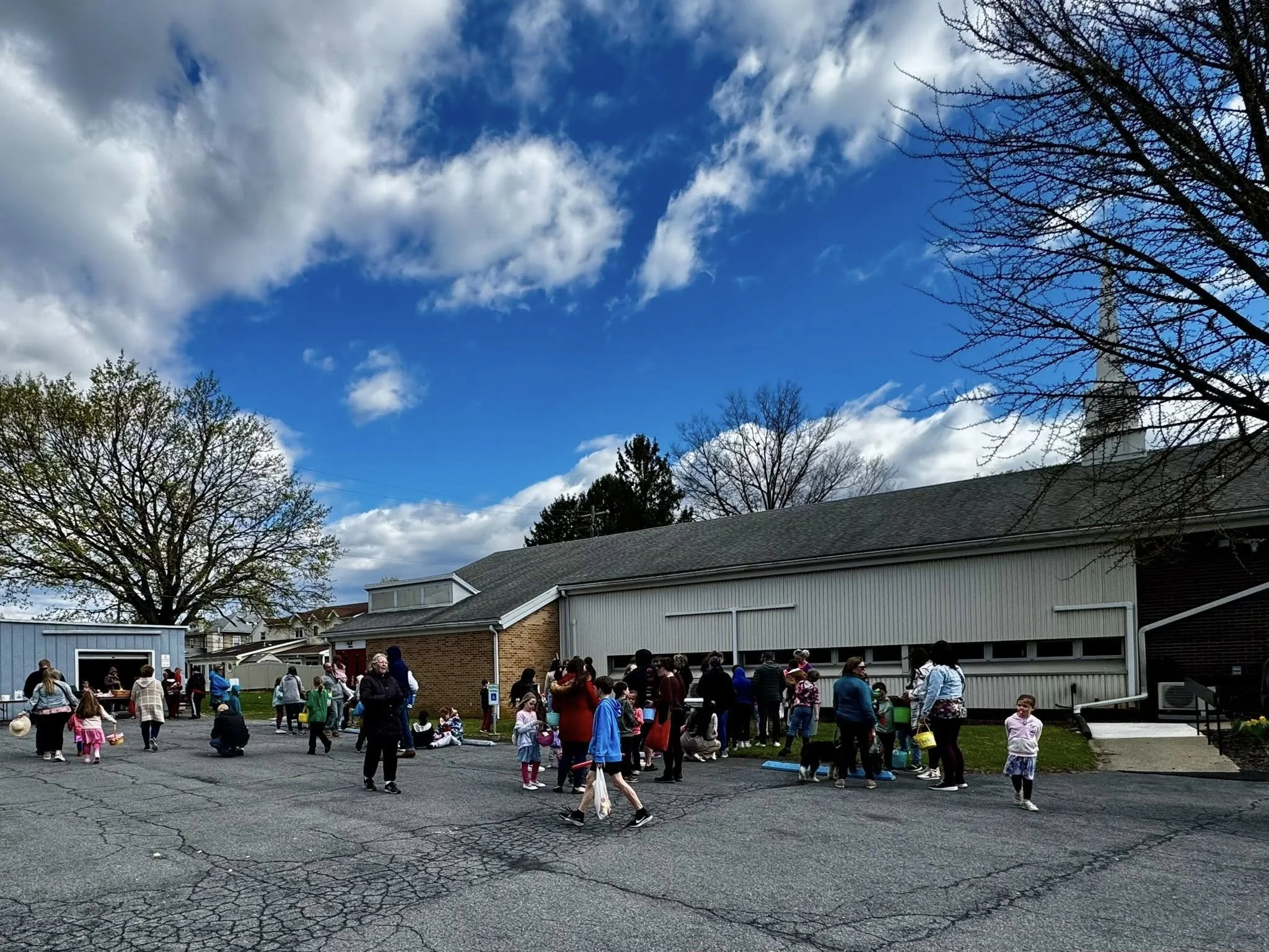 People gathered outside a building with a grey roof, some holding yellow buckets, and children sitting on the ground; trees with some budding leaves and clouds in the sky.