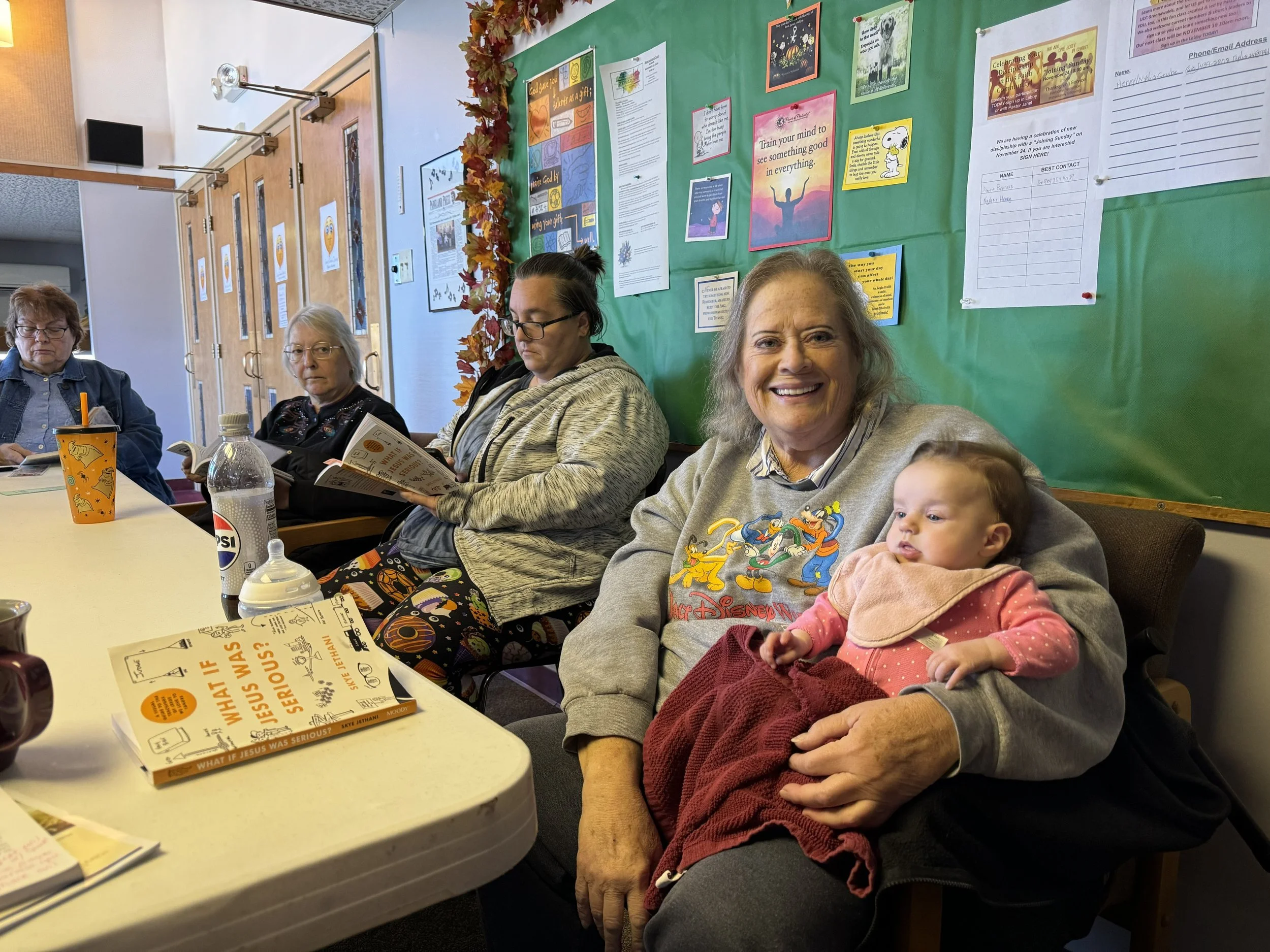 Four adults and one baby sitting at a table in a room with a green bulletin board on the wall behind them. The bulletin board has various posters and notices, one with a sunset and a person with arms raised, and another with a Snoopy drawing. The adults appear to be looking at books, papers, or their phones. The baby is sitting on the woman in the beige sweatshirt, wearing a pink sweatshirt and red pants, and looking to the right.