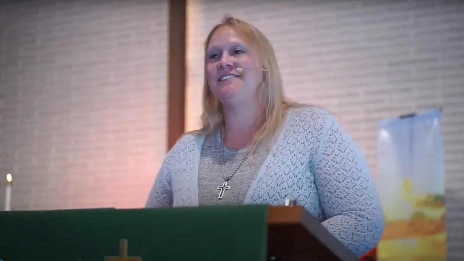 A woman with long blonde hair wearing a gray top, white crocheted cardigan, and a cross necklace, standing or sitting behind a closed green church lectern with a gold cross, inside a church or similar setting.