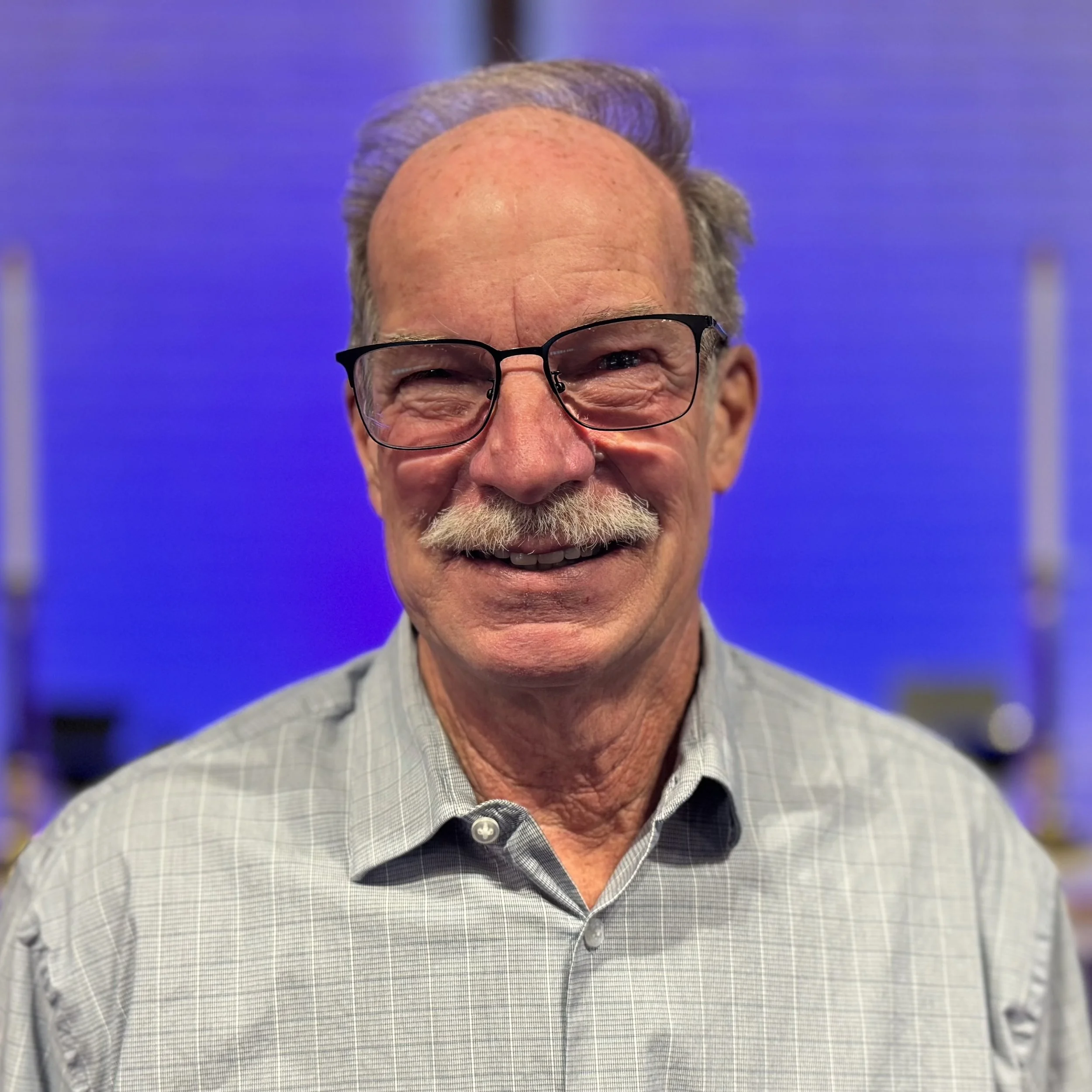 Close-up of an elderly man wearing glasses and a checkered shirt smiling, with a blurred blue background.
