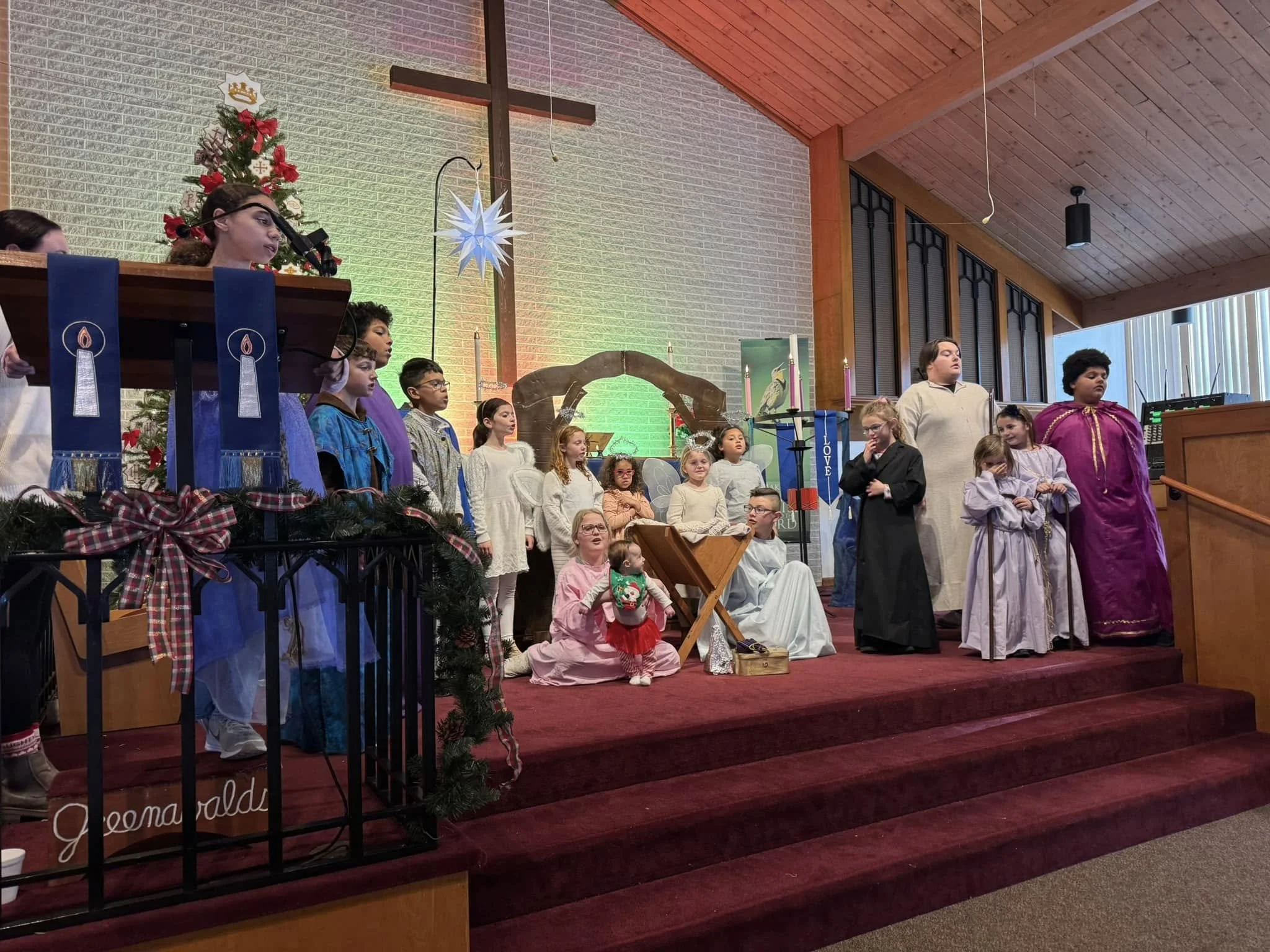 Children dressed in costumes participating in a Christmas nativity play on stage inside a church decorated with a Christmas tree, ornaments, and candles.