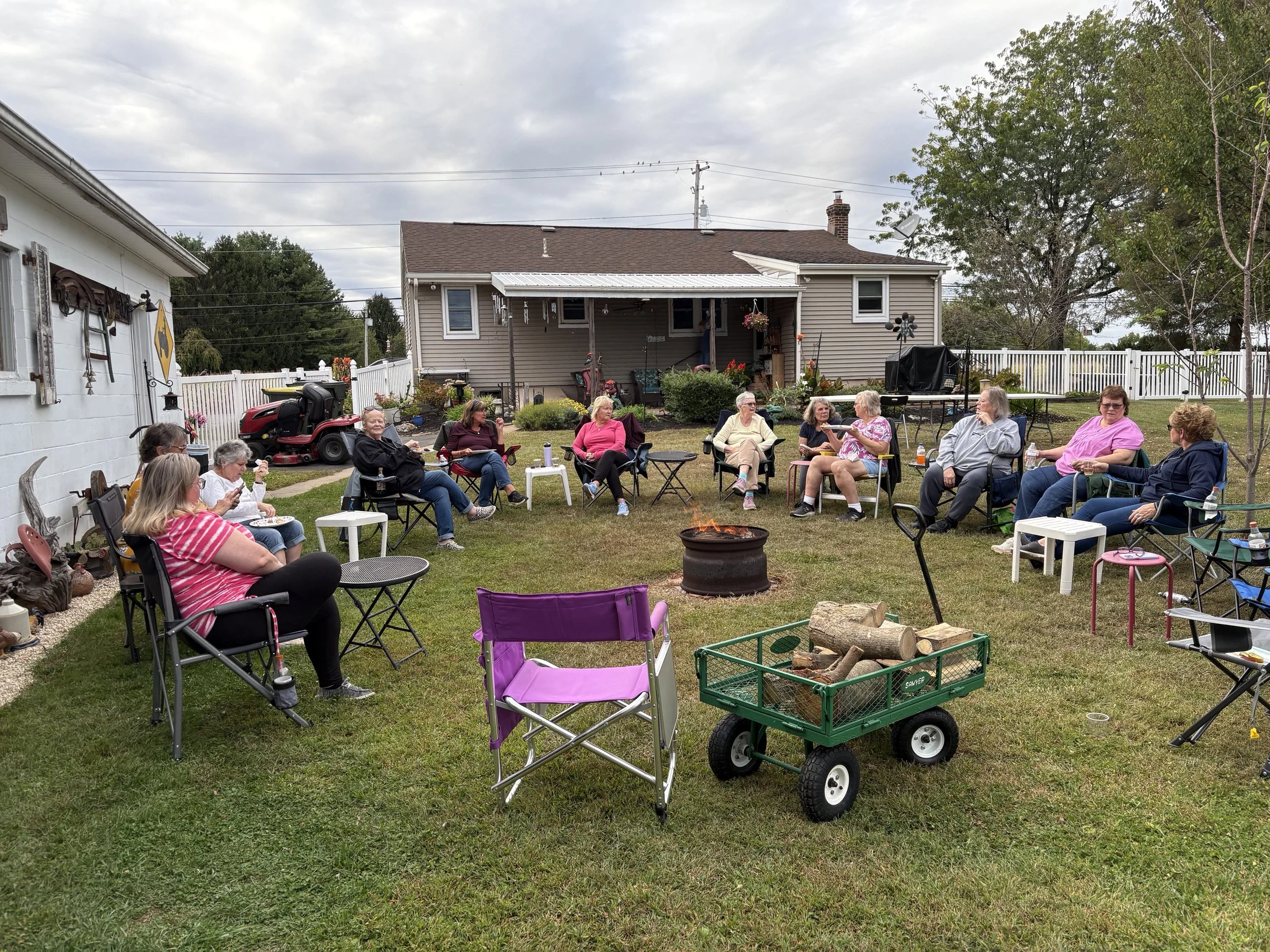 A group of people sitting in lawn chairs around a fire pit in a backyard during daytime. There are houses, trees, and a white fence in the background.