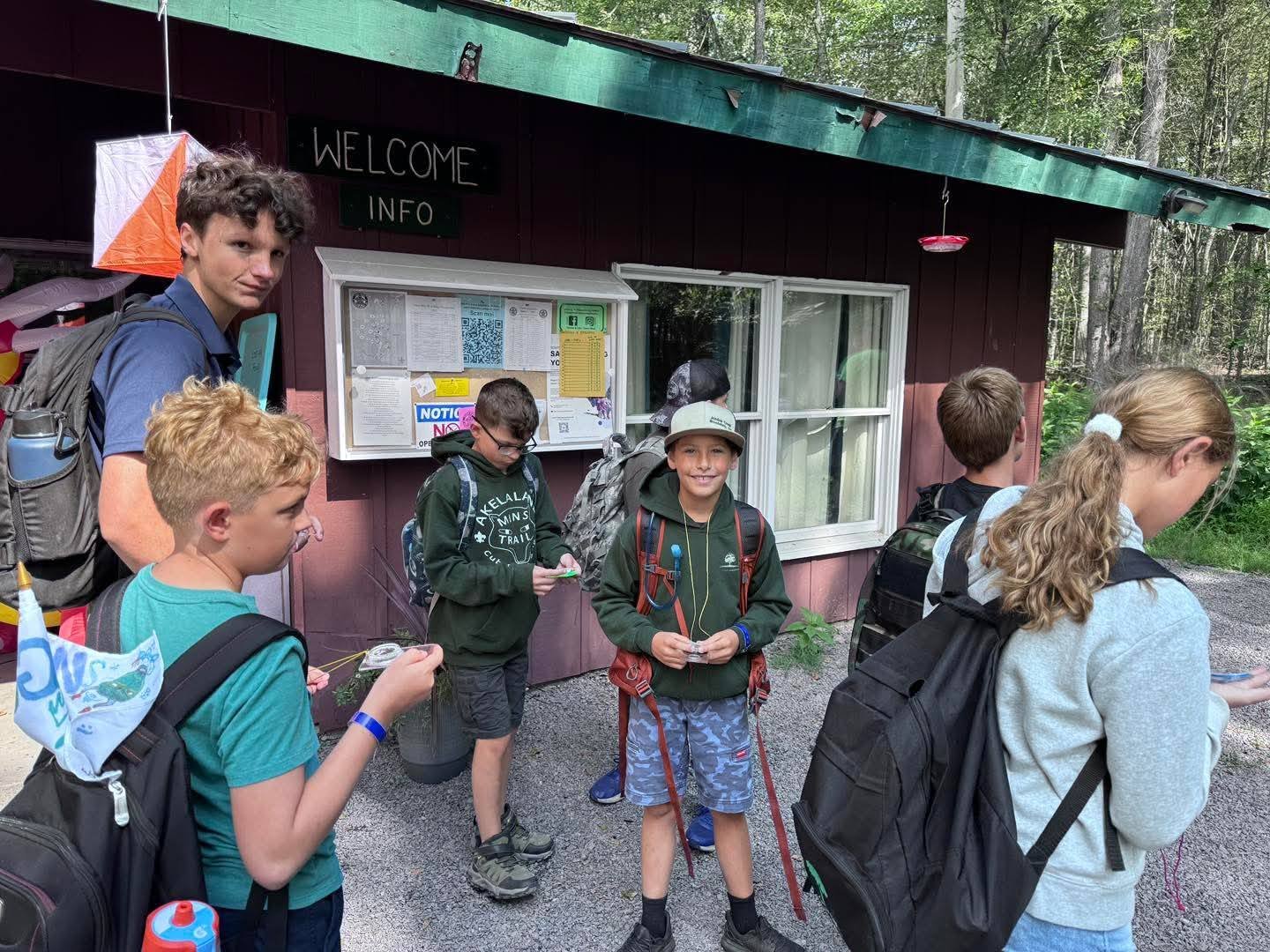 Group of children and a teenage guide standing outside a wilderness visitor center, some children holding electronics or keys, all with backpacks, near a bulletin board on a cabin-style building in a forested area.