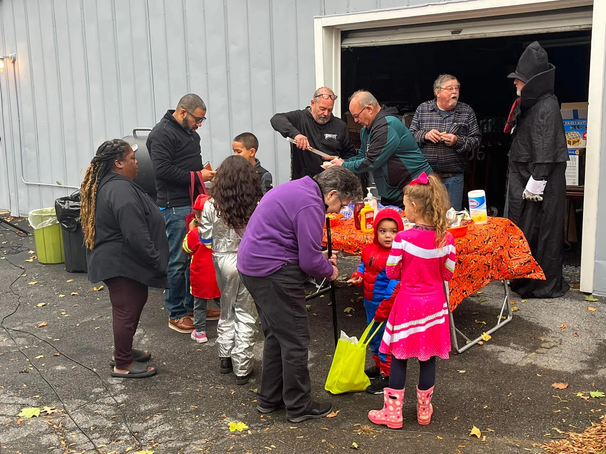 A group of children and adults gather around a table outside a garage, enjoying Halloween treats. The children are wearing colorful costumes including a pink dress with rain boots and a red jacket, while adults are dressed casually, some with contain