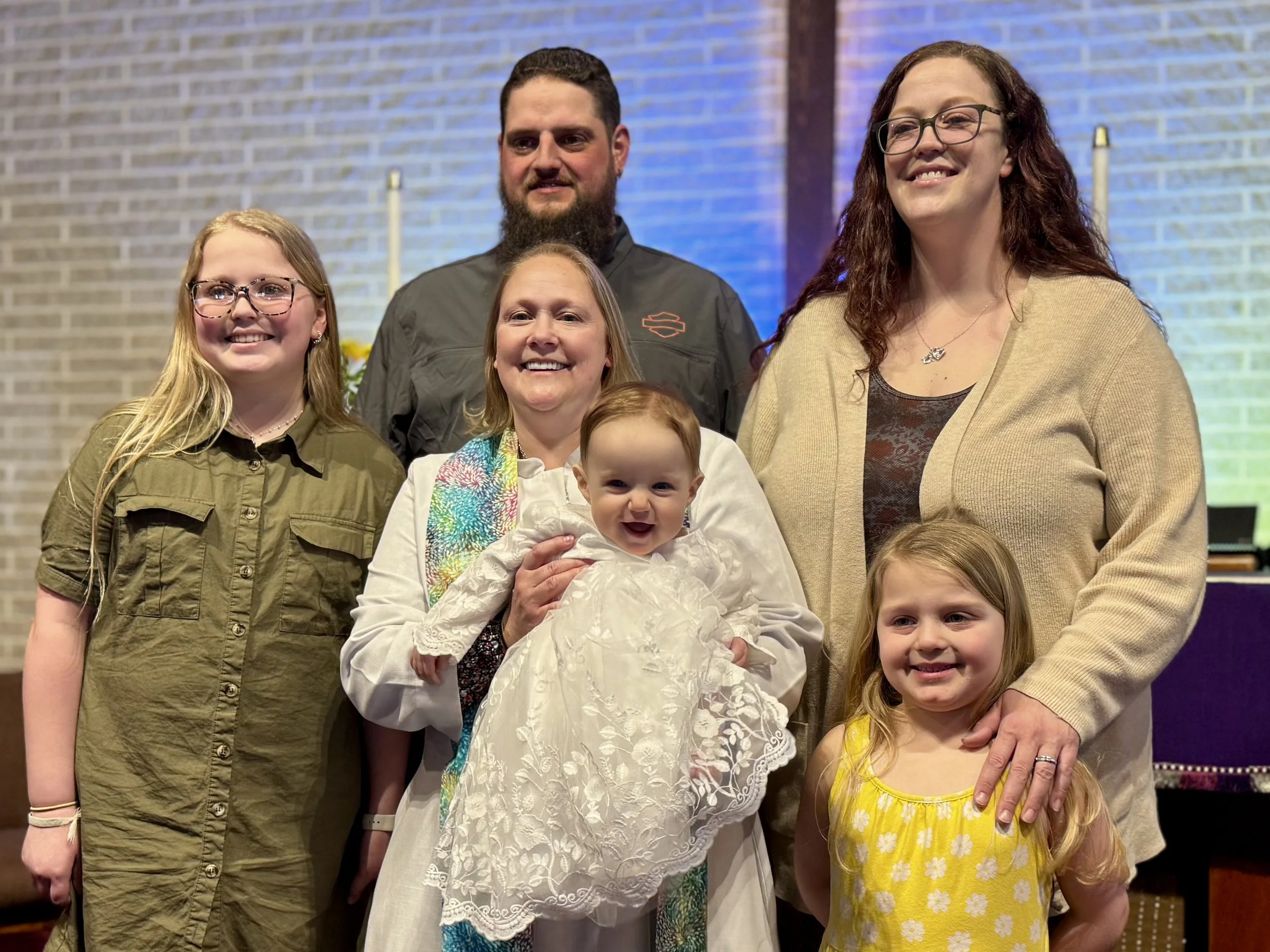 A group of seven people standing together in front of a brick wall, smiling, including a woman holding a baby. The group consists of children and adults with diverse appearances.