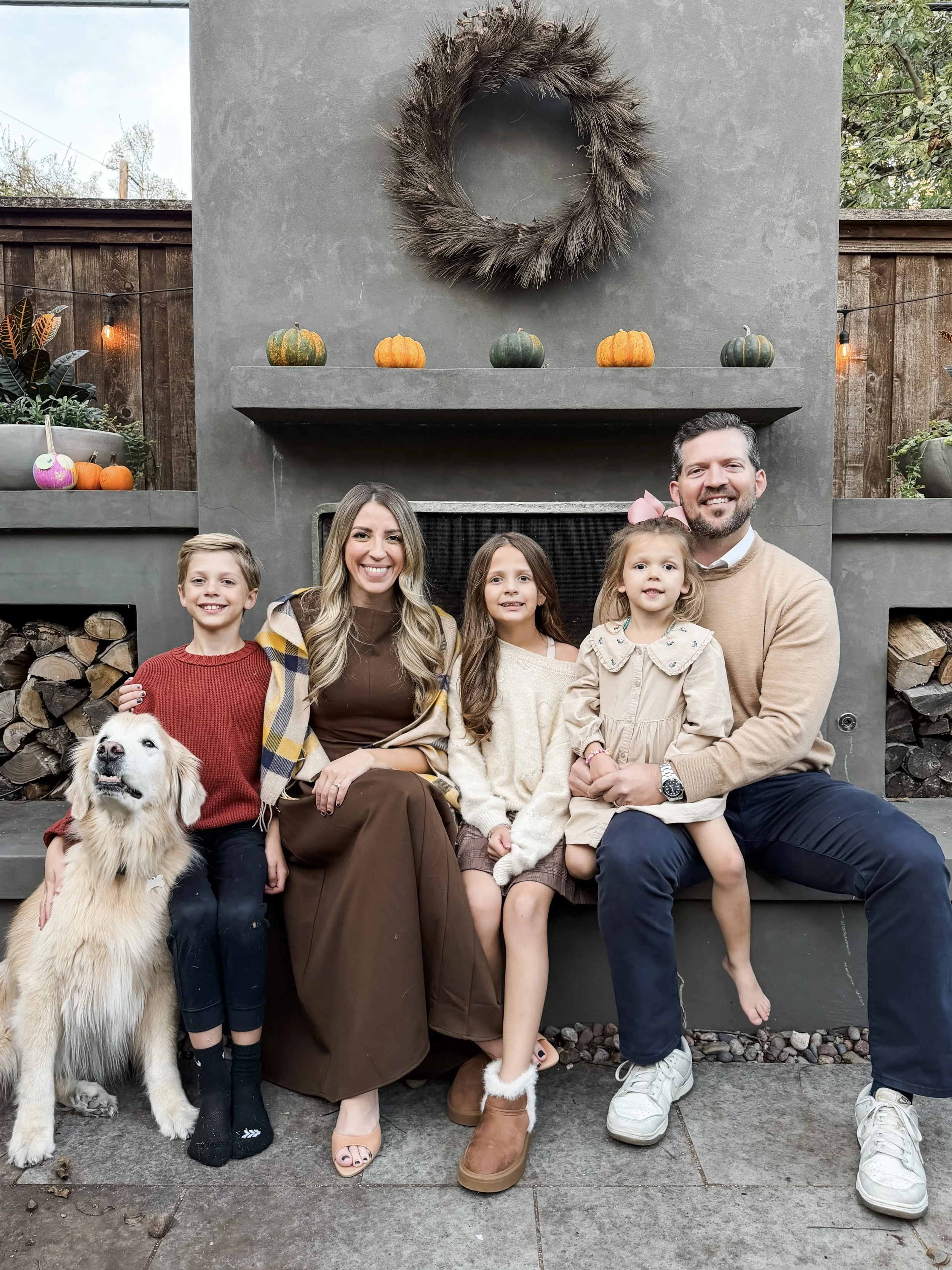 Family of five and dog sitting on a concrete patio in front of a modern fireplace with pumpkins and fall decorations, including a large wreath hanging above.