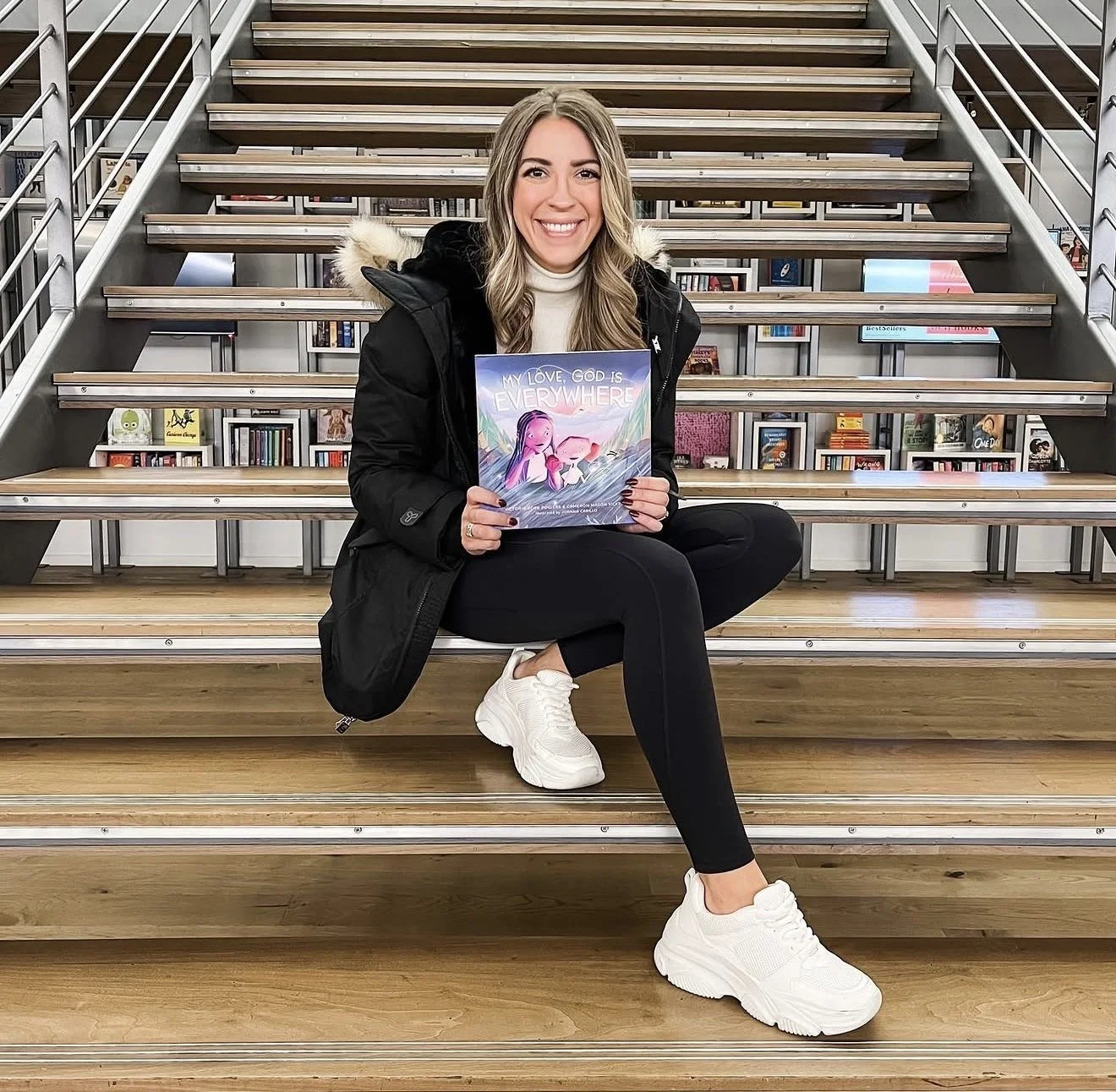 A young woman with blonde hair, wearing a black jacket, white turtleneck, and athletic shoes, sitting on a wooden staircase in a bookstore, holding a colorful book titled "My Love, God is Everywhere," smiling at the camera.