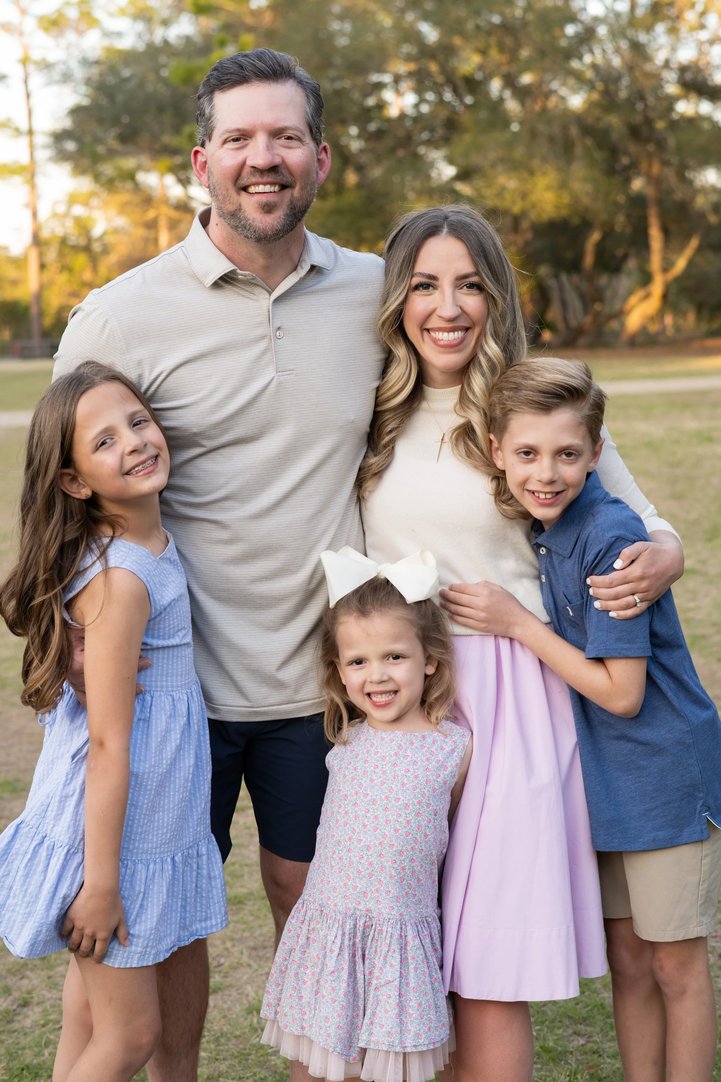 A happy family of five standing outdoors on a grassy field with trees in the background during daylight. The family members include two adults and three children, all smiling and dressed in casual clothing.