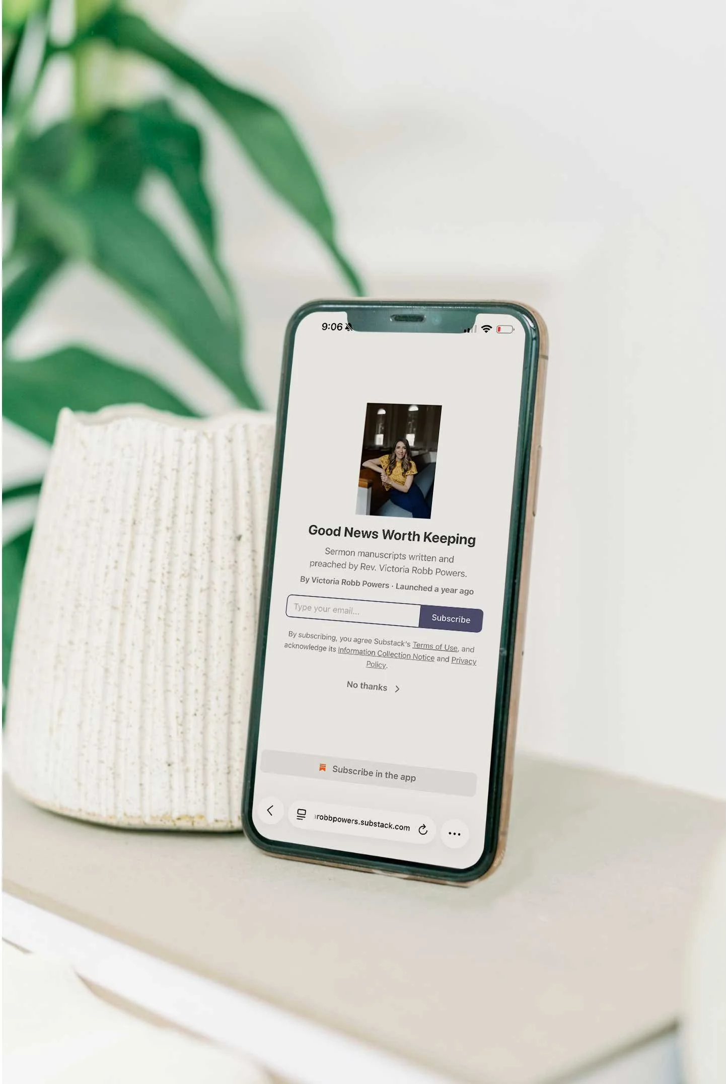 Close-up of a smartphone showing a subscription webpage for sermon manuscripts, placed on a white surface next to a white ceramic pot with a green leafy plant in the background.