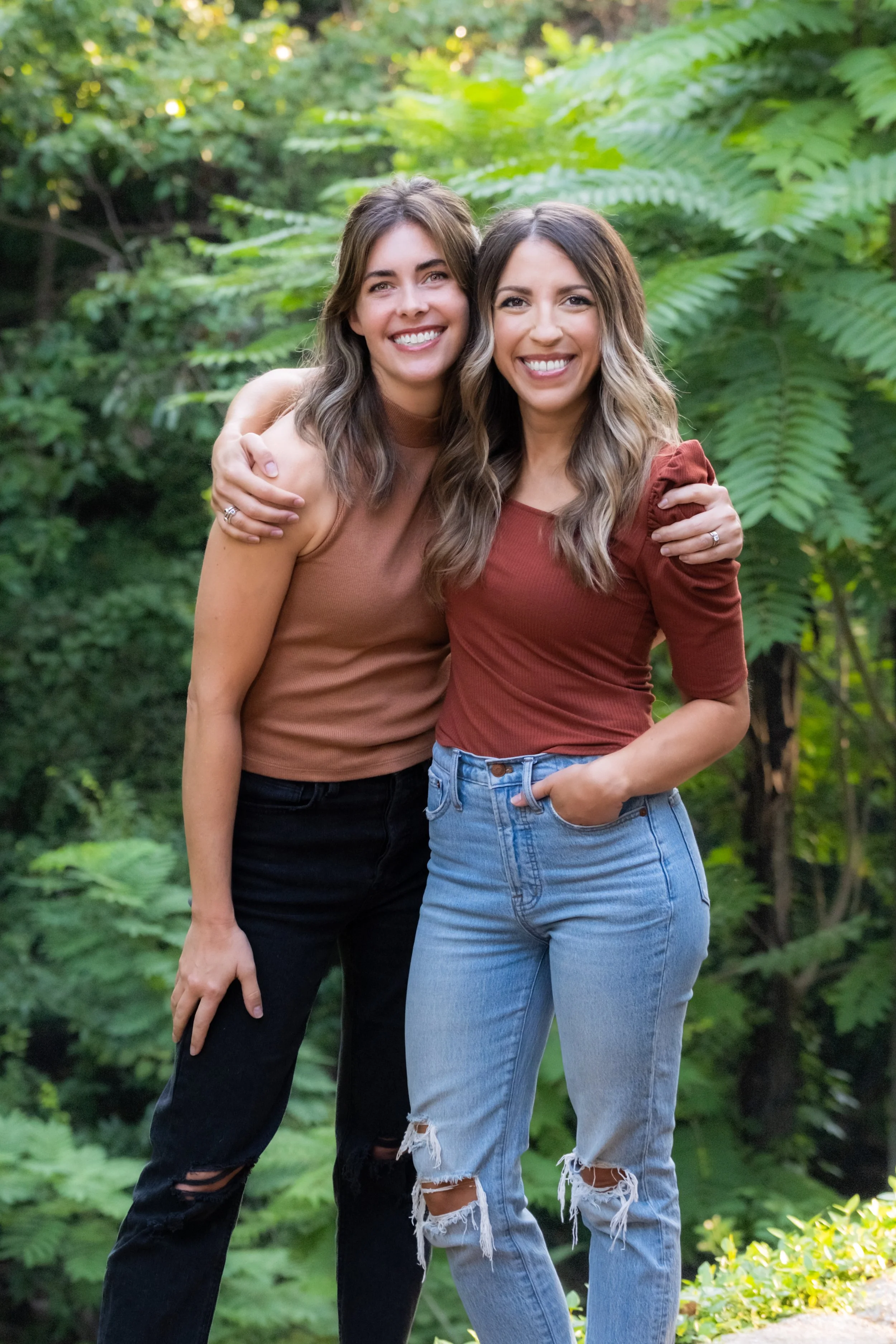 Two young women smiling and hugging outdoors in front of lush green plants.