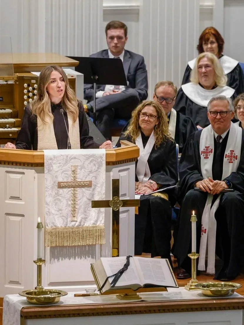 A woman speaking at a church pulpit during a religious service, surrounded by clergy and church officials seated behind her.