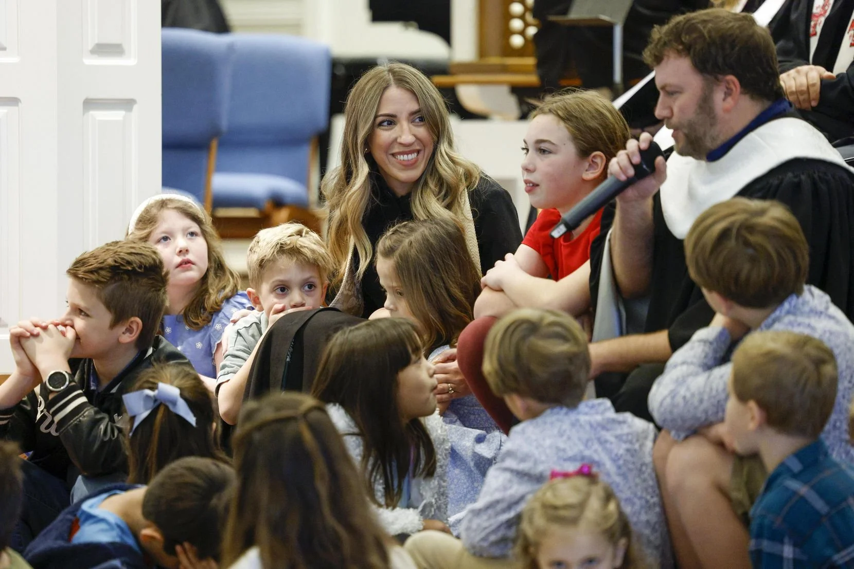 A woman and children sitting on the floor during a gathering, with a man dressed in clergy robes holding a microphone and speaking.