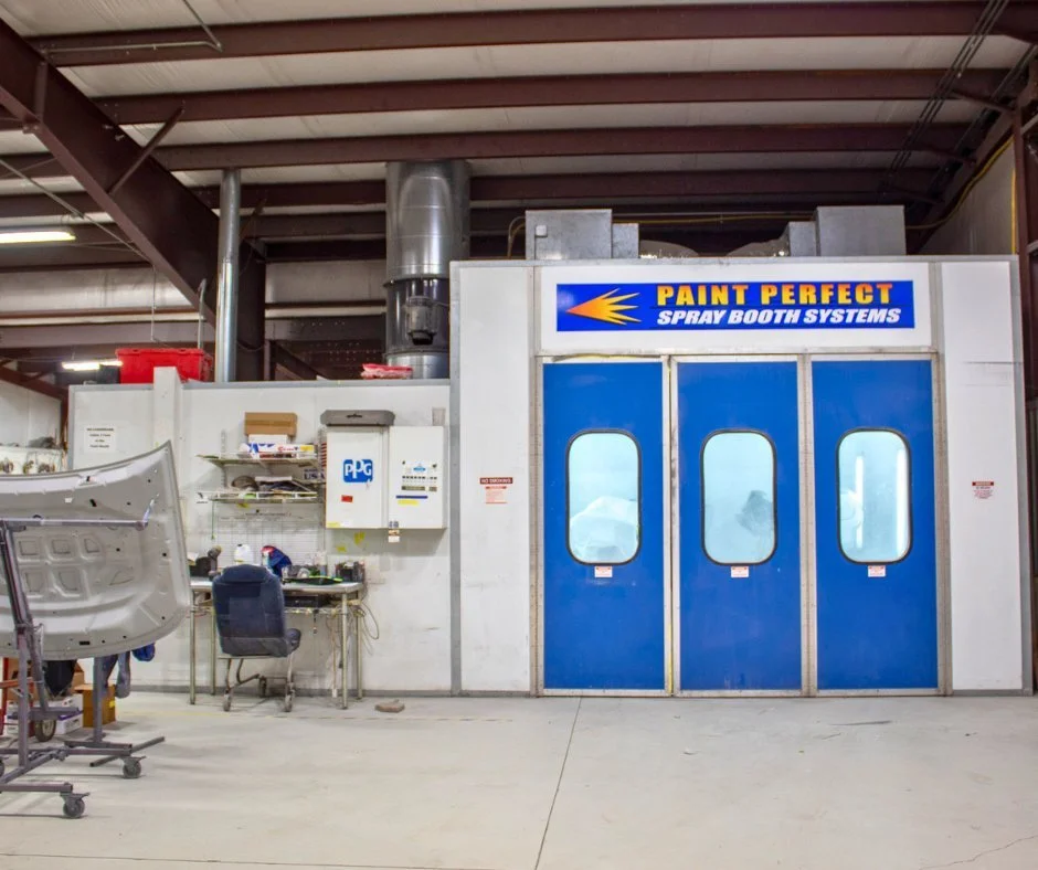A spray booth system labeled 'Paint Perfect Spray Booth Systems' in a workshop or industrial setting, with a large blue and white enclosed spray booth and a workstation with tools and a chair nearby.