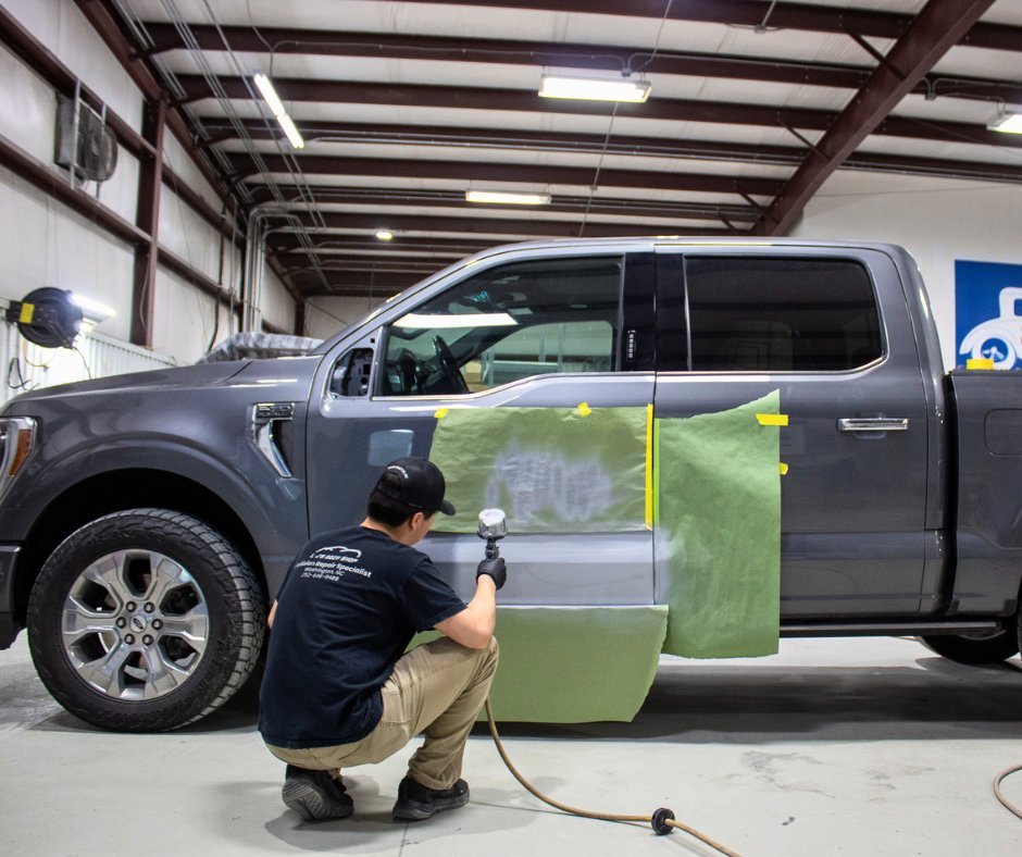 A technician in a black shirt and khaki pants is applying a protective film to the side of a gray pickup truck inside a workshop. The worker is kneeling and using a spray gun on the vehicle's door area, with green masking paper partially covering the truck.
