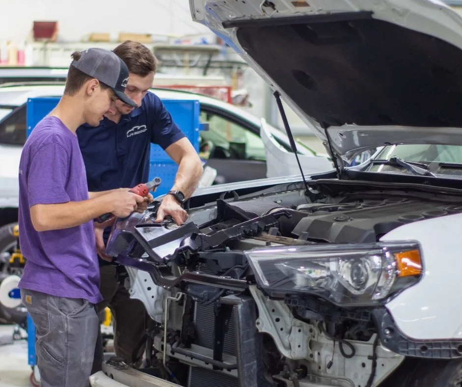 Two men working on a white car with its hood open in an auto repair shop.