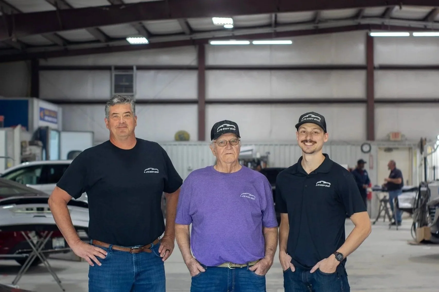 Three men standing inside a garage or workshop, wearing matching black caps and T-shirts, with a car partially visible behind them. The man in the middle is wearing a purple T-shirt, while the other two are in black. They are posing and smiling at the camera.