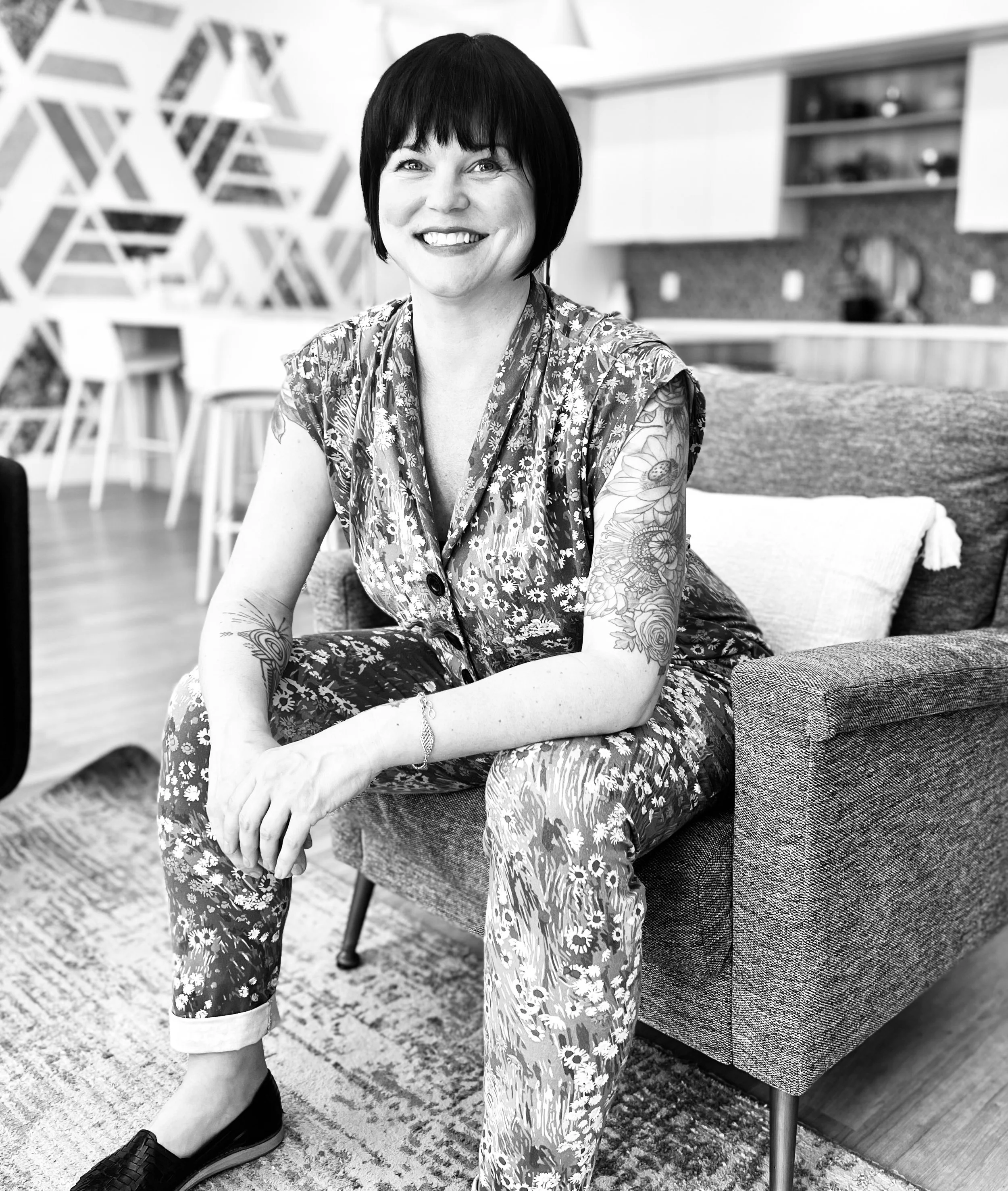A woman with short dark hair, smiling, wearing a floral patterned sleeveless dress, sitting on a sofa in a modern living room with patterned wallpaper and wooden floor.
