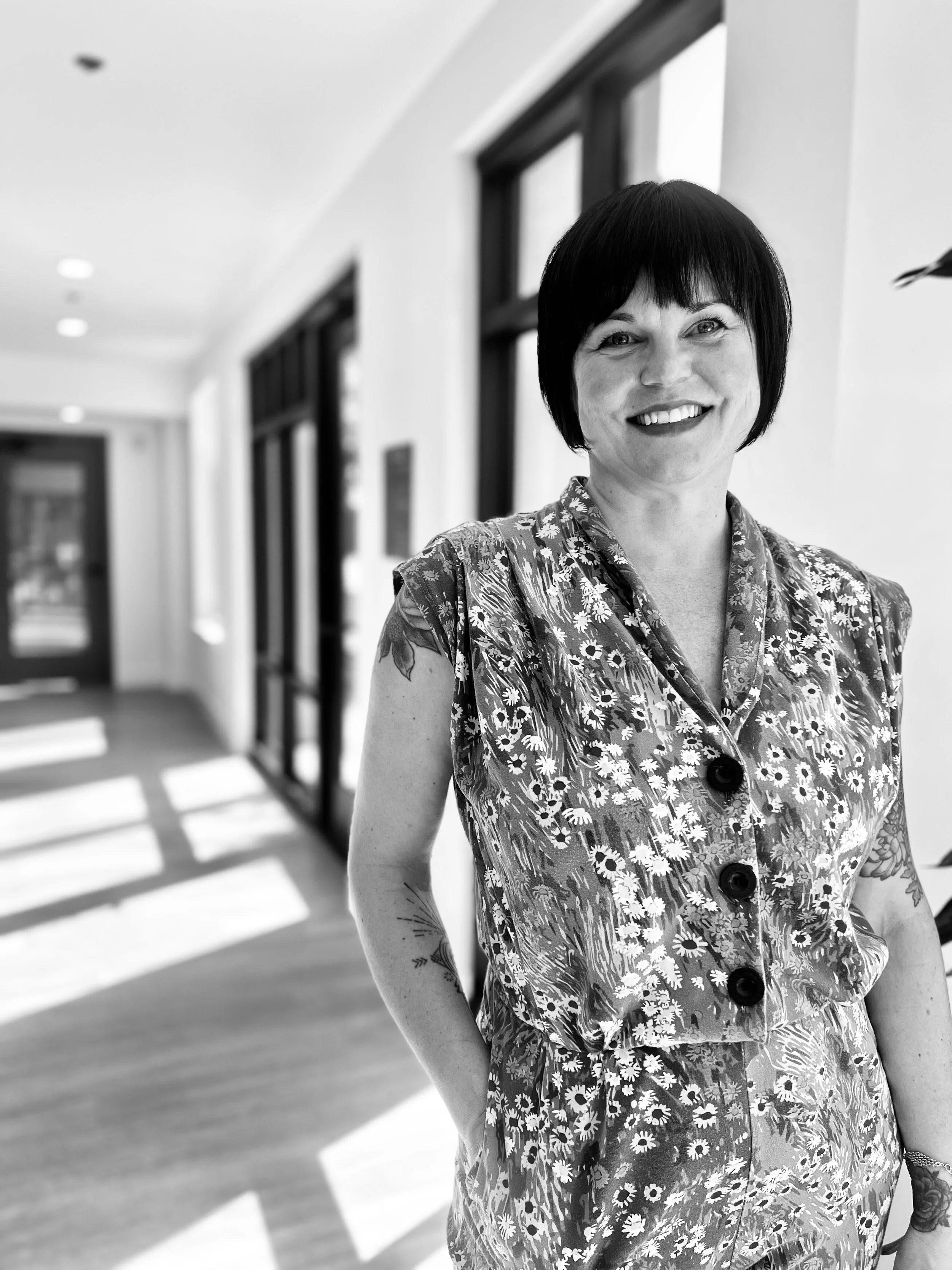 A woman with short black hair smiling, wearing a floral patterned sleeveless dress, standing in a brightly lit room with large windows and blinds.