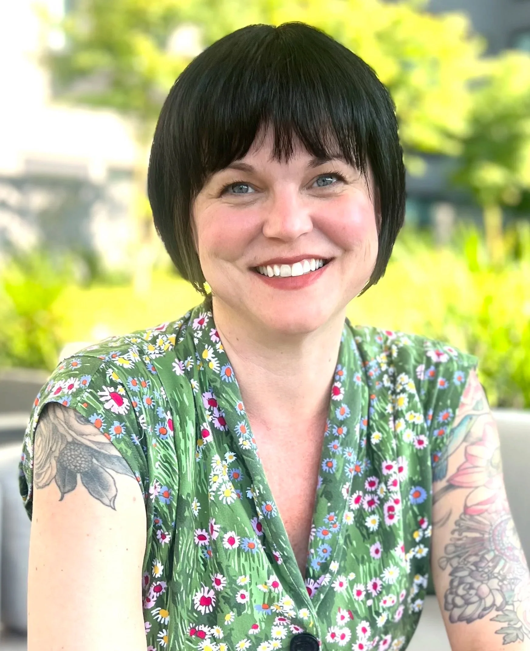 Close-up of a woman with short black hair, blue eyes, and tattoos on her arm, smiling outdoors against a blurred green background. She wears a green dress with a colorful floral pattern.