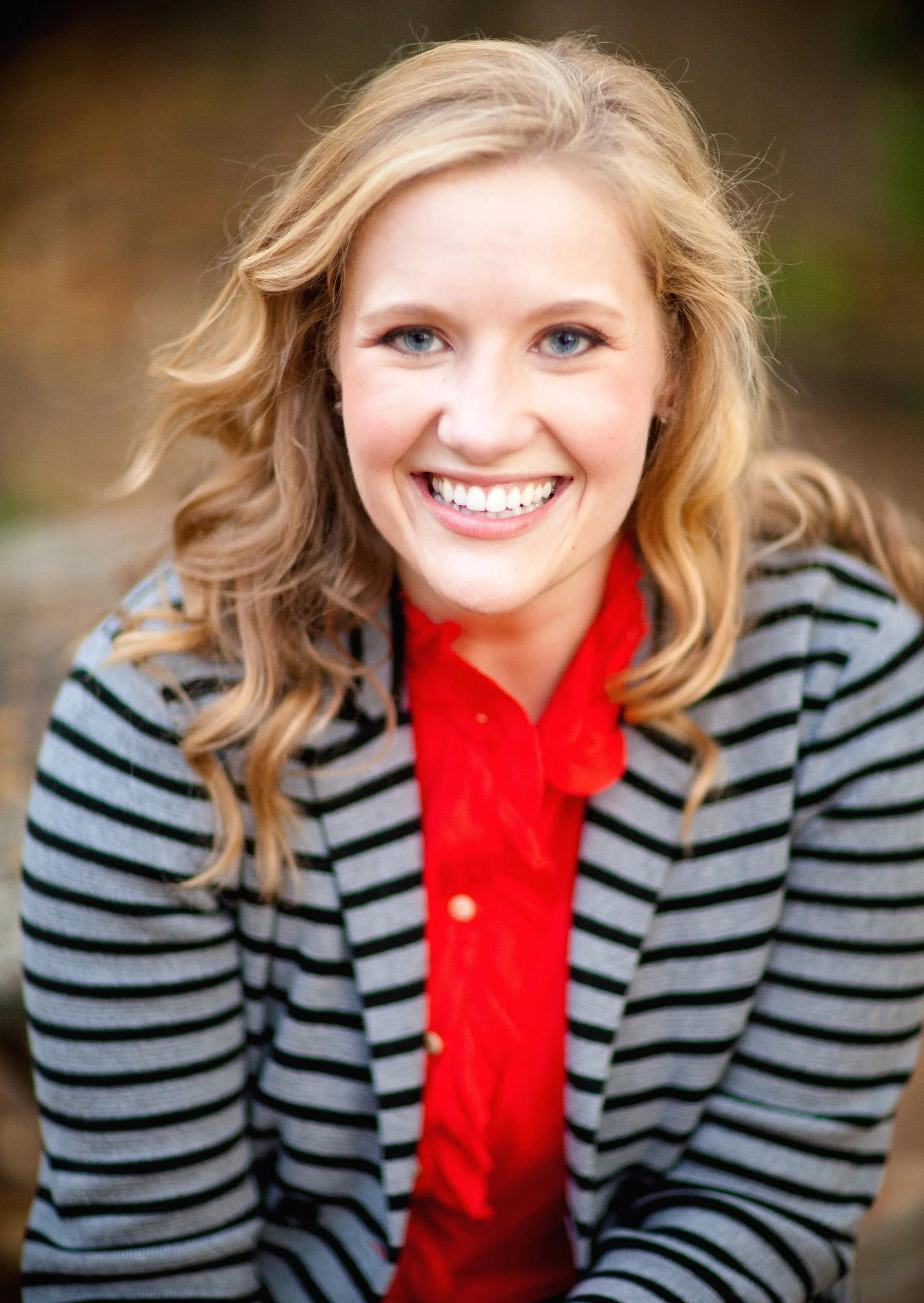 Close-up portrait of a smiling woman with blonde hair, wearing a striped blazer and red shirt, outdoors.