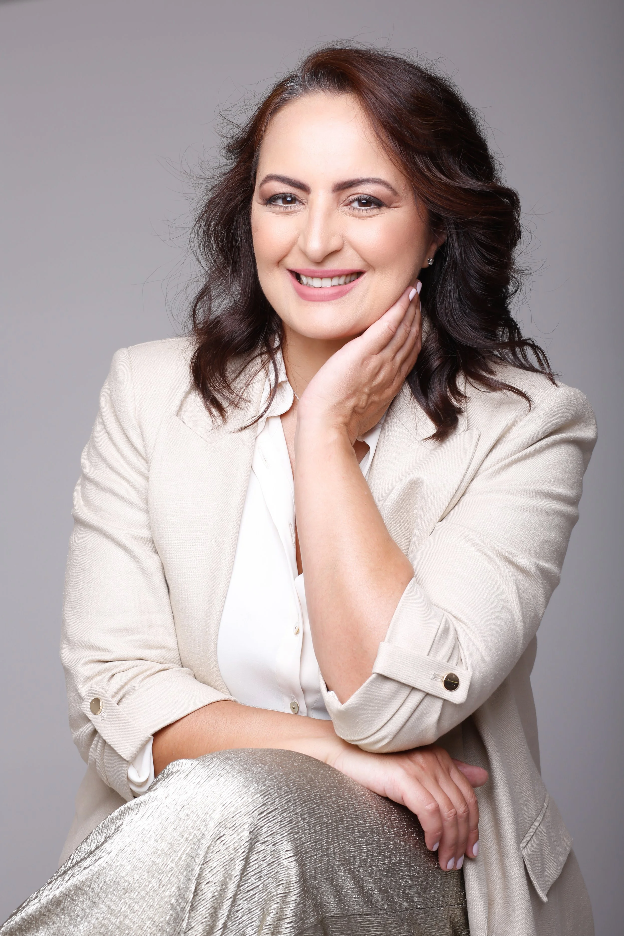 A smiling woman with dark brown wavy hair, wearing a light-colored blazer and shimmering pants, sitting with her hand on her chin against a plain gray background.