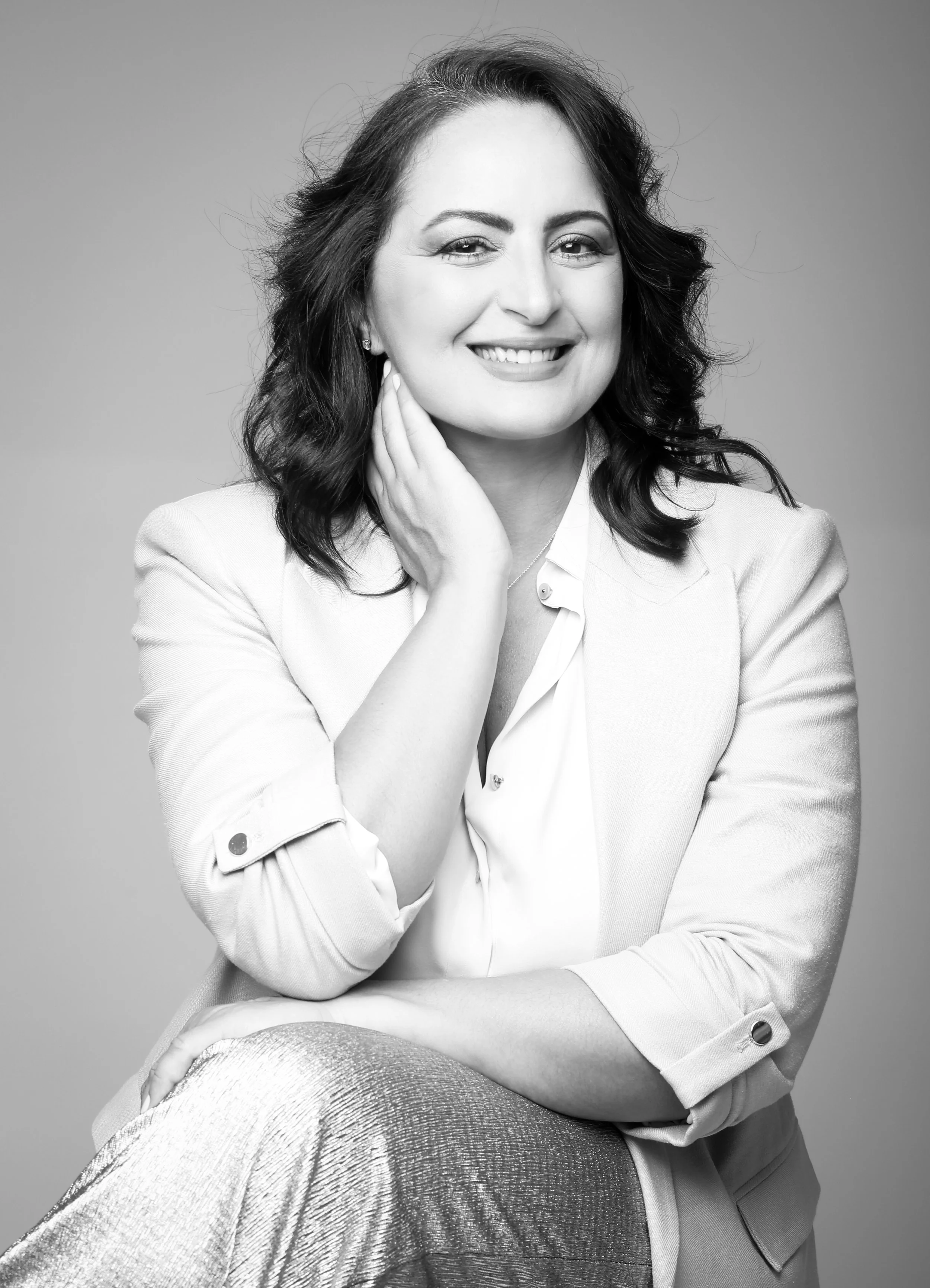 Black and white portrait of a woman smiling, with her hand resting on her neck, wearing a blazer and a blouse, posed against a plain background.