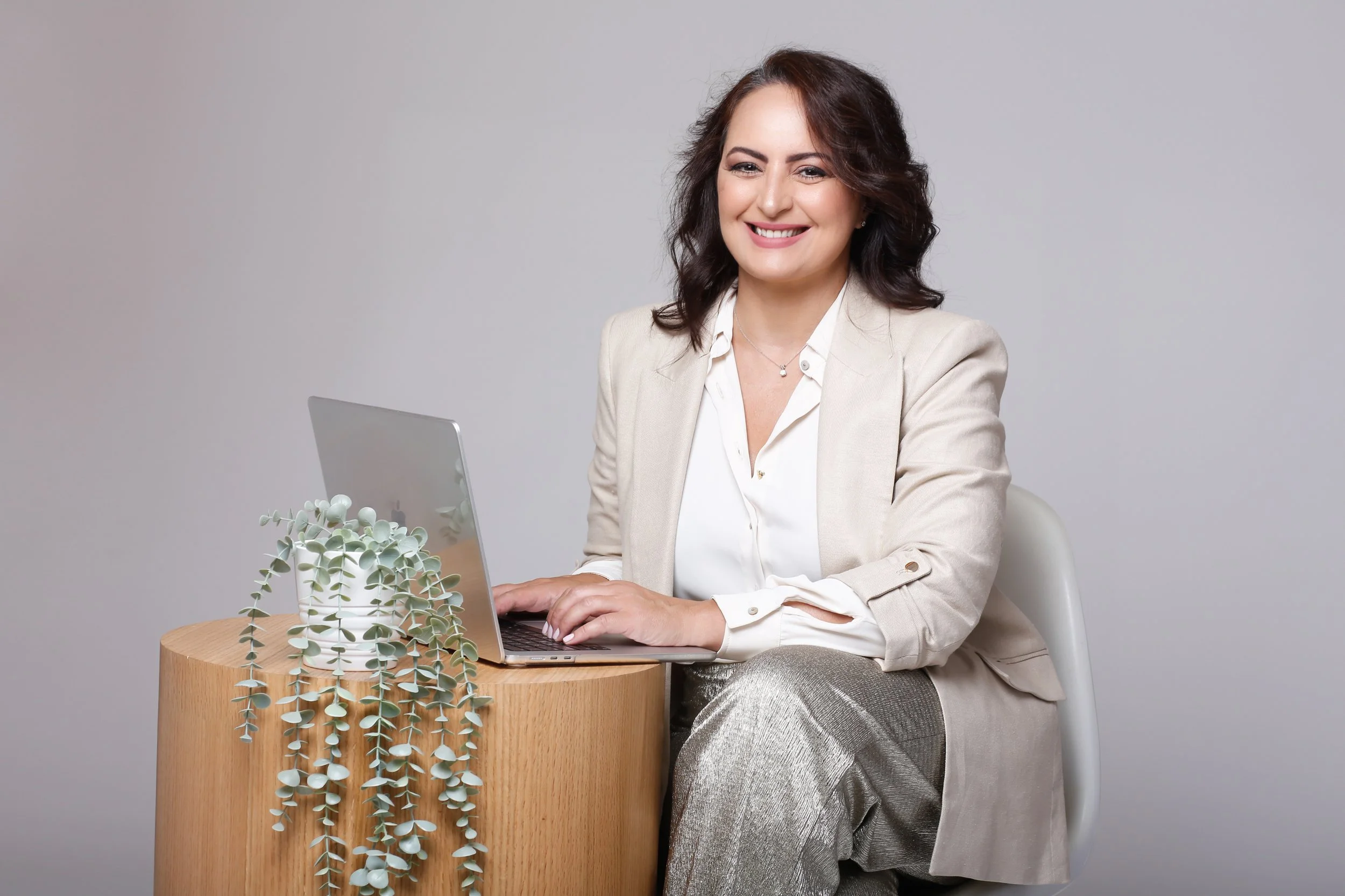 A smiling woman with dark brown wavy hair, sitting at a round wood table, working on a silver laptop. She is dressed in a beige blazer and a white blouse, with a small necklace. There is a white potted plant with green leaves on the table, against a plain gray background.