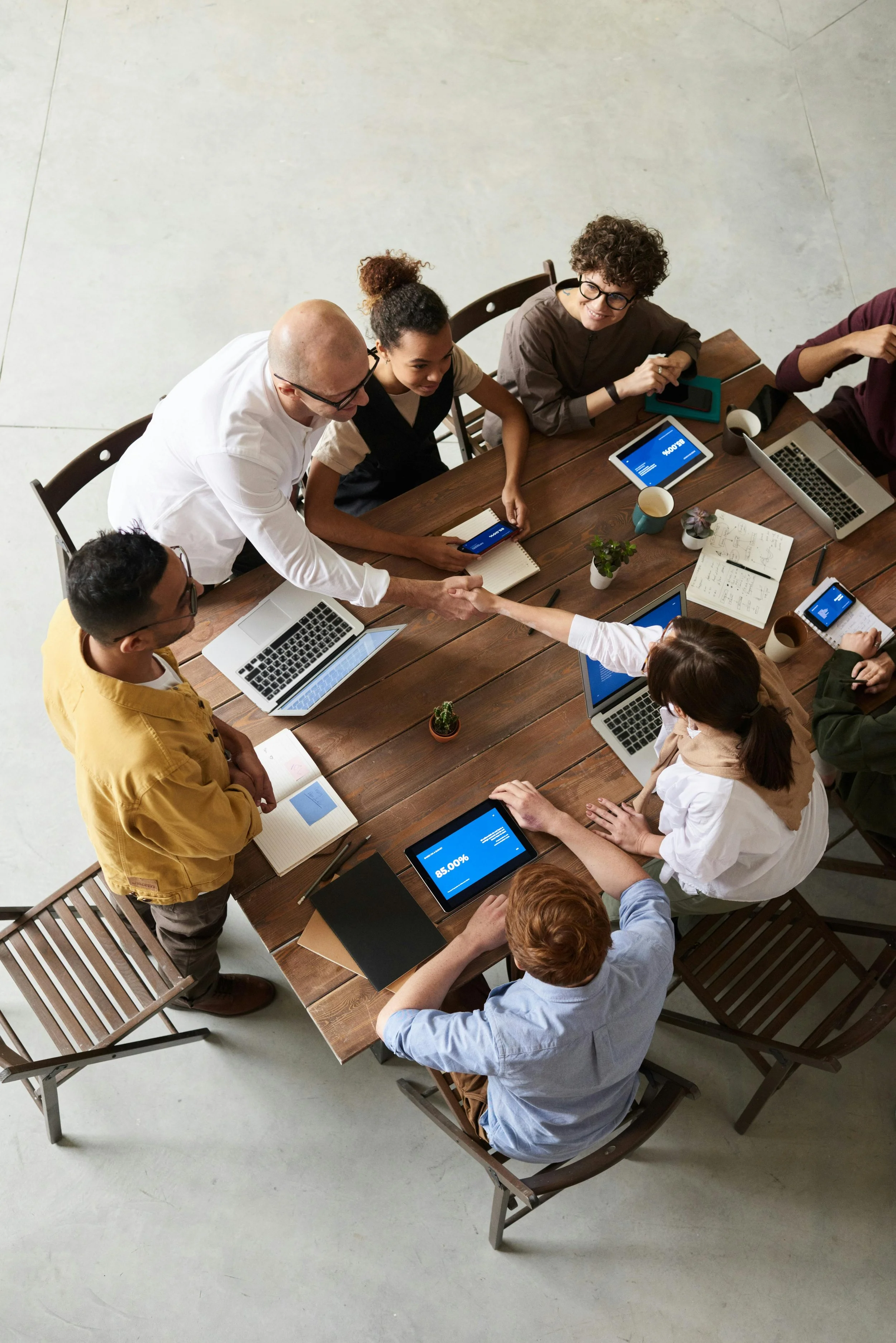 A diverse group of eight people gathered around a large wooden table in a modern office, engaged in a business meeting. They are using laptops, tablets, and smartphones, with some shaking hands, and one person is taking notes. The table has plants, notebooks, and coffee cups, and the floor is light-colored concrete.