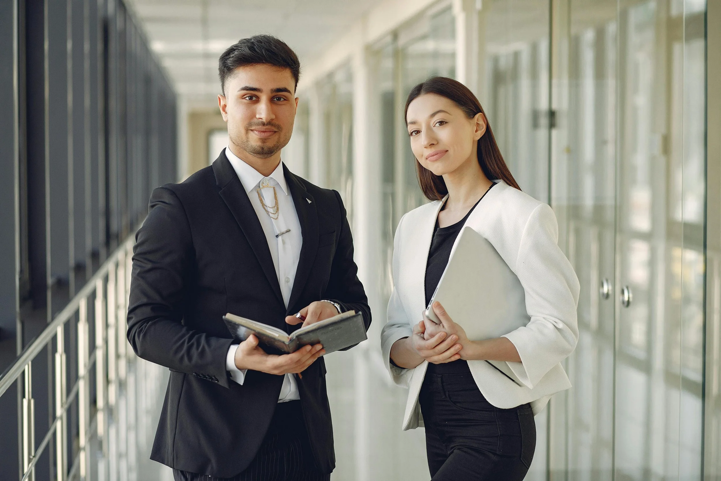A man in a black suit and a woman in a white blazer standing in a modern office corridor with glass walls. They are looking at the camera, and the man is holding a notebook while the woman is holding a folder or tablet.
