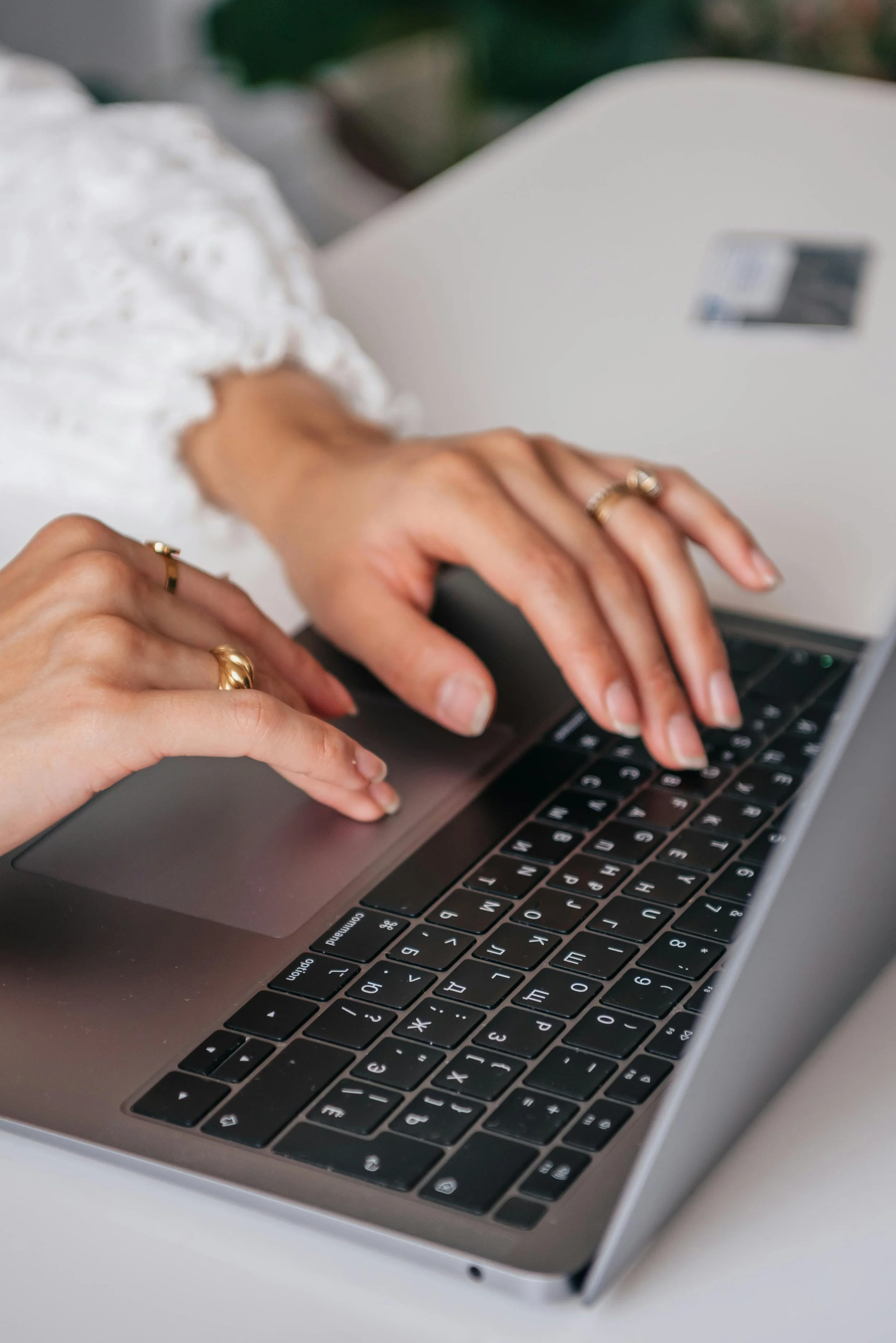 Close-up of a person's hands typing on a laptop keyboard. The person is wearing rings and a white, ruffled sleeve. The laptop has a sleek, silver body with black keys.