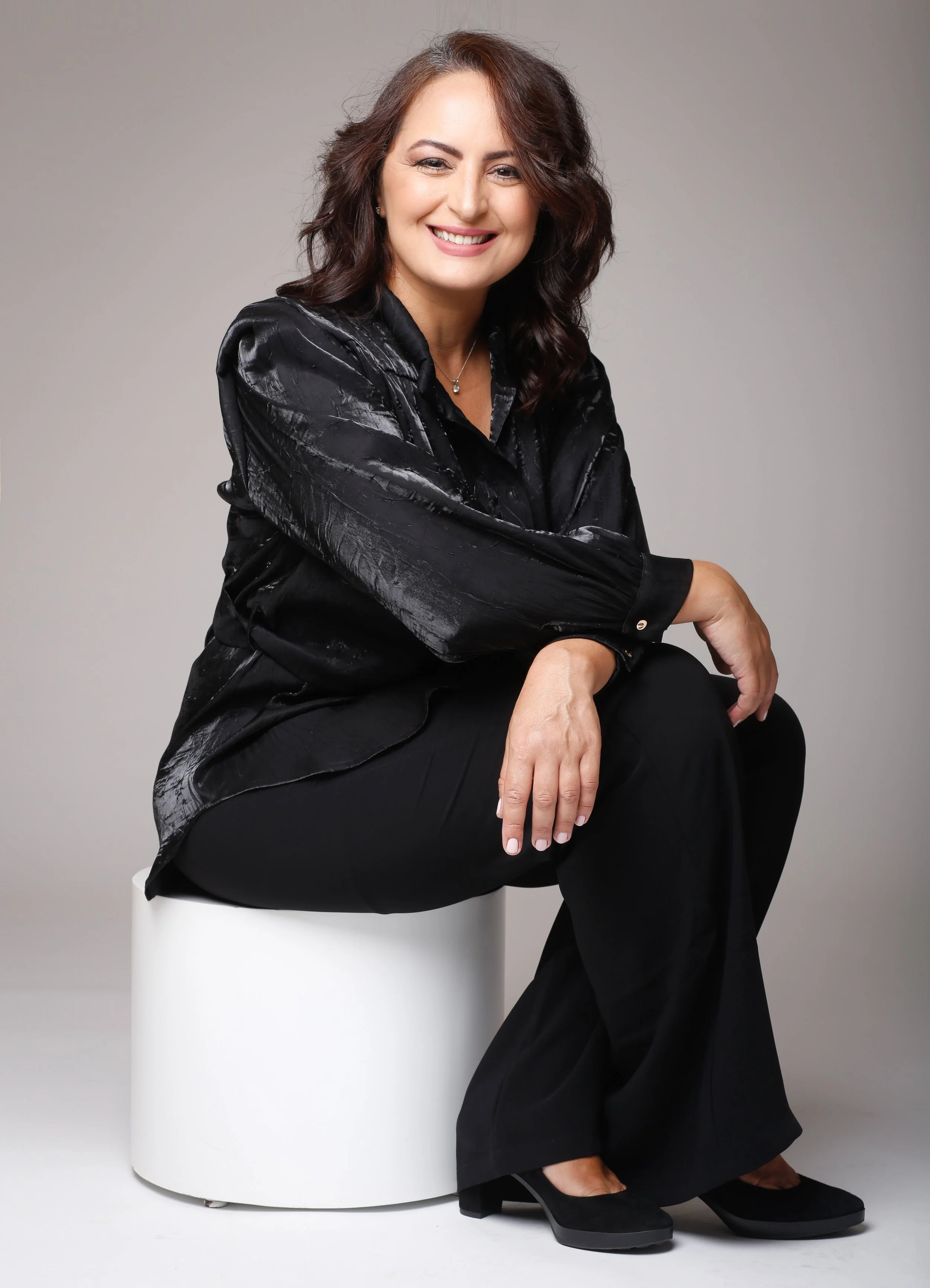 A woman sitting on a white stool, wearing a black shiny blouse and black wide-legged trousers, with black shoes, smiling and posing against a plain gray background.